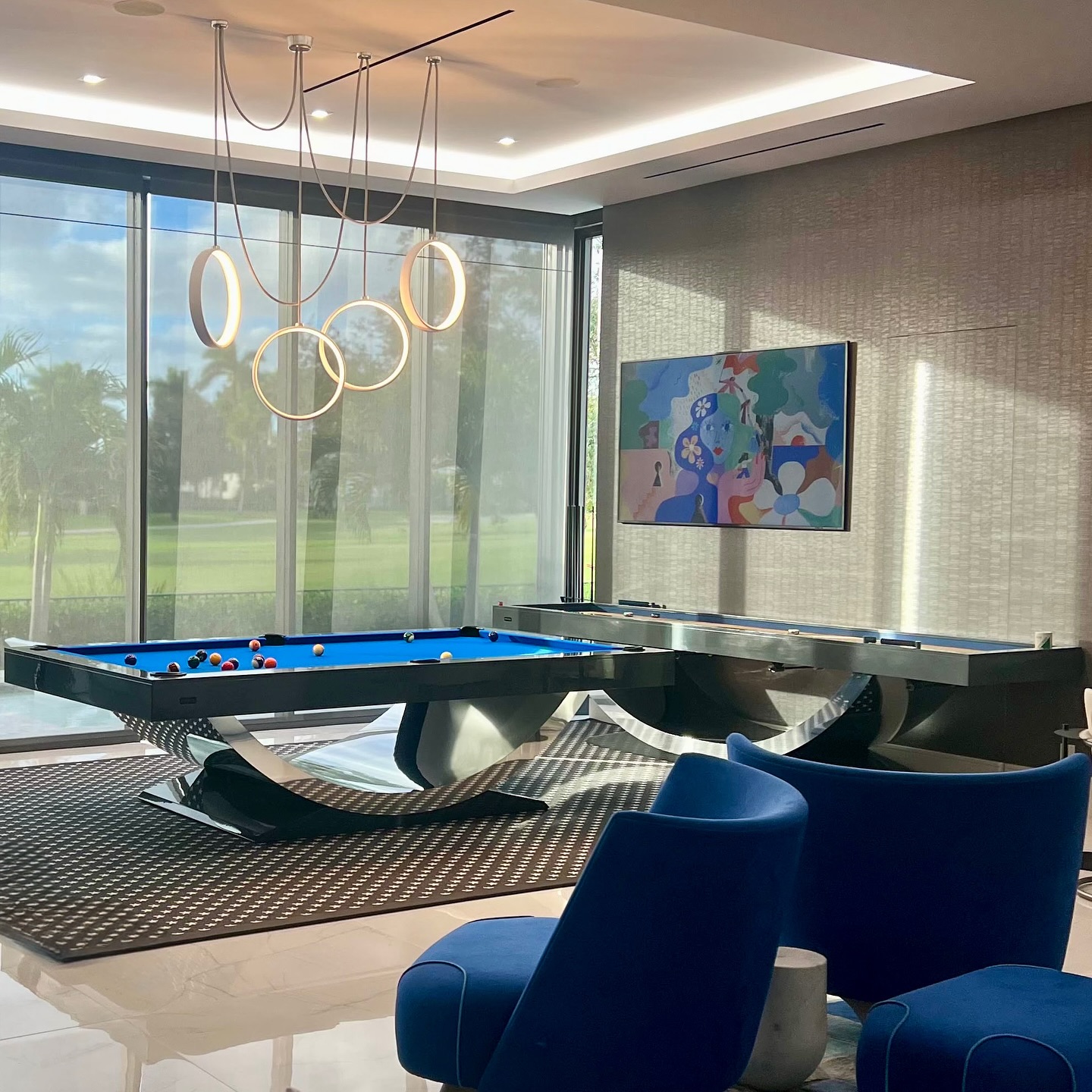 A modern room with a blue pool table, abstract wall art, two blue chairs, and large windows letting in natural light. A unique chandelier hangs from the ceiling, and greenery is visible outside.