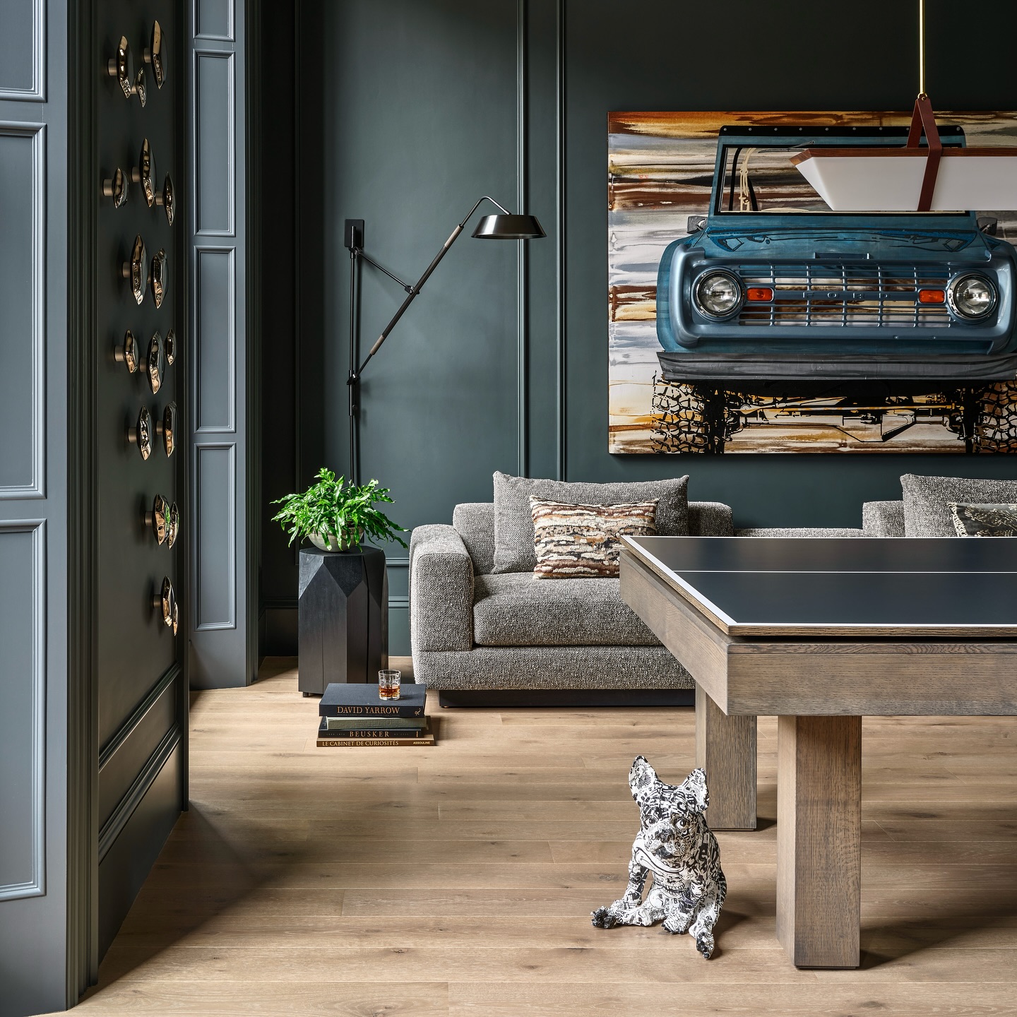 A modern living room with gray walls, a sofa, a wall-mounted lamp, a large painting of a blue vintage truck, and a pool table. A decorative dog statue sits on a wooden floor next to plants and books.