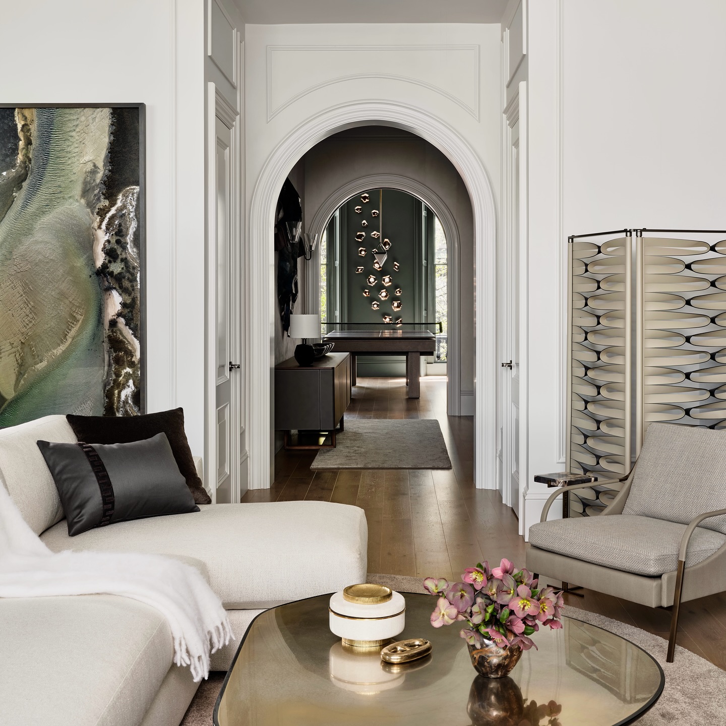 A modern living room with a curved white sofa, armchair, abstract wall art, and a gold coffee table with flowers. An arched doorway leads to a room with a pool table and hanging pendant lights.