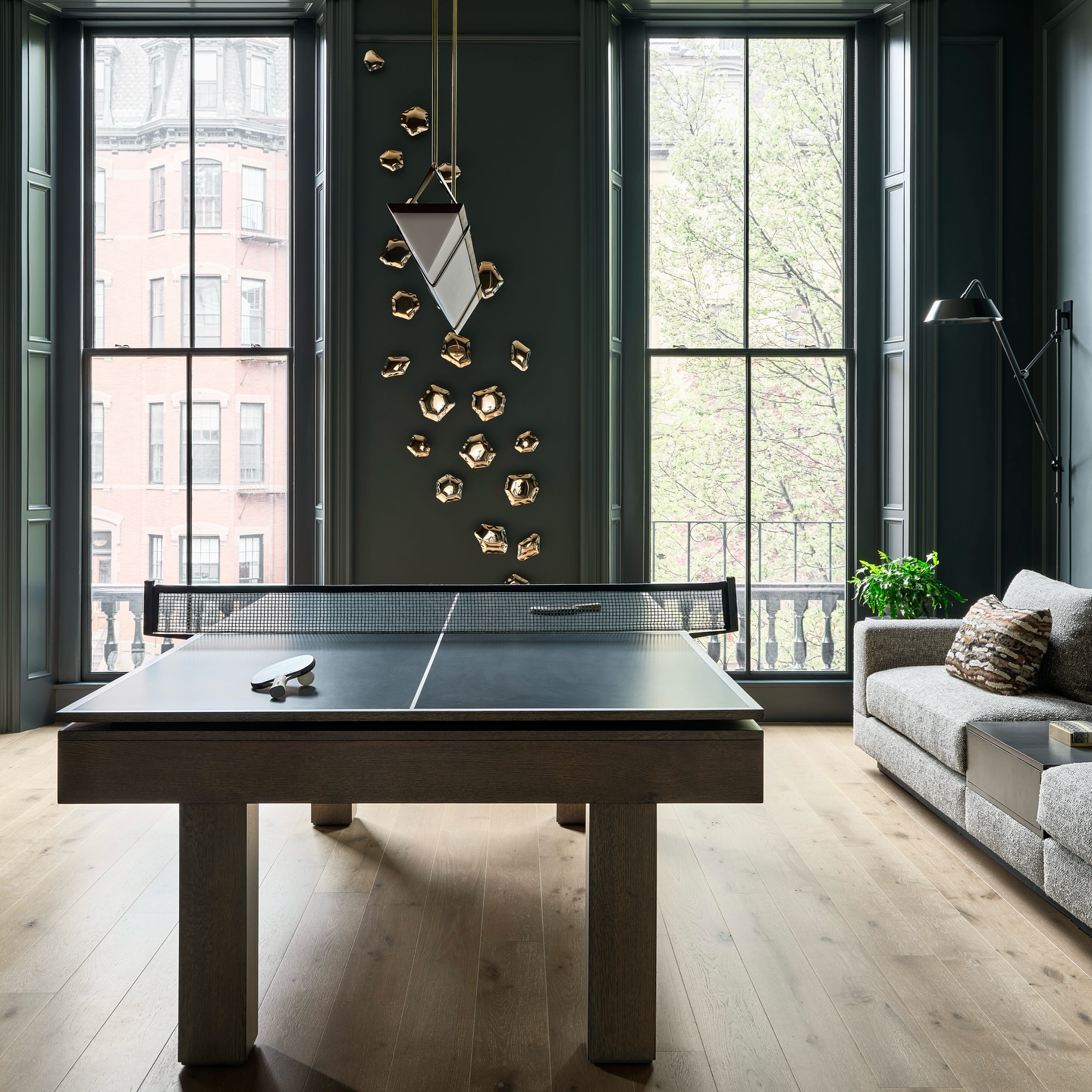 A modern living room with tall windows, a black ping pong table, a gray sofa, wall art, a geometric pendant light, and a view of trees and buildings outside.