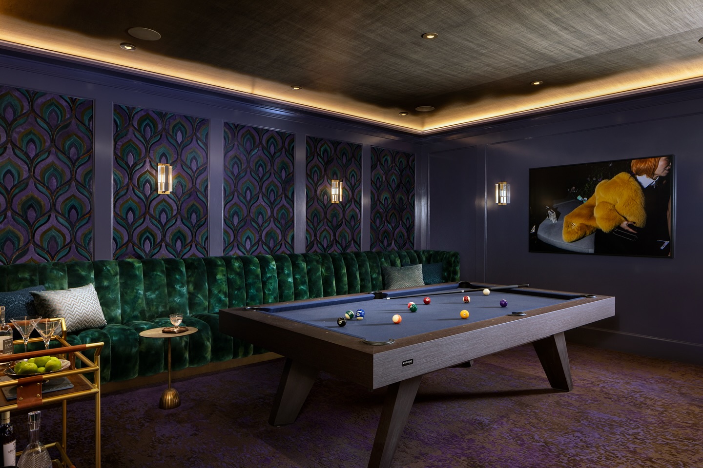 A stylish game room with a modern pool table, green velvet tufted bench, art deco wallpaper, wall-mounted lights, a TV screen, and a gold bar cart with drinks and fruit.