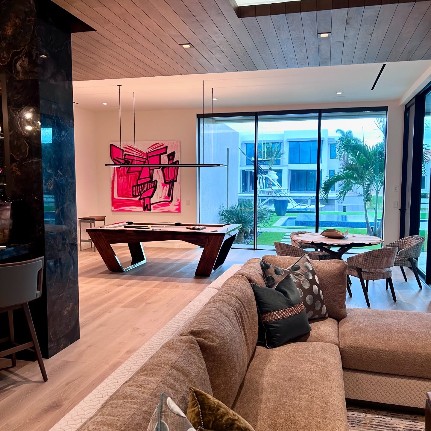 A modern living room with a brown sectional sofa, a pool table, and a dining area. Large windows show a view of palm trees and a contemporary building. A bold pink abstract painting hangs on the wall.
