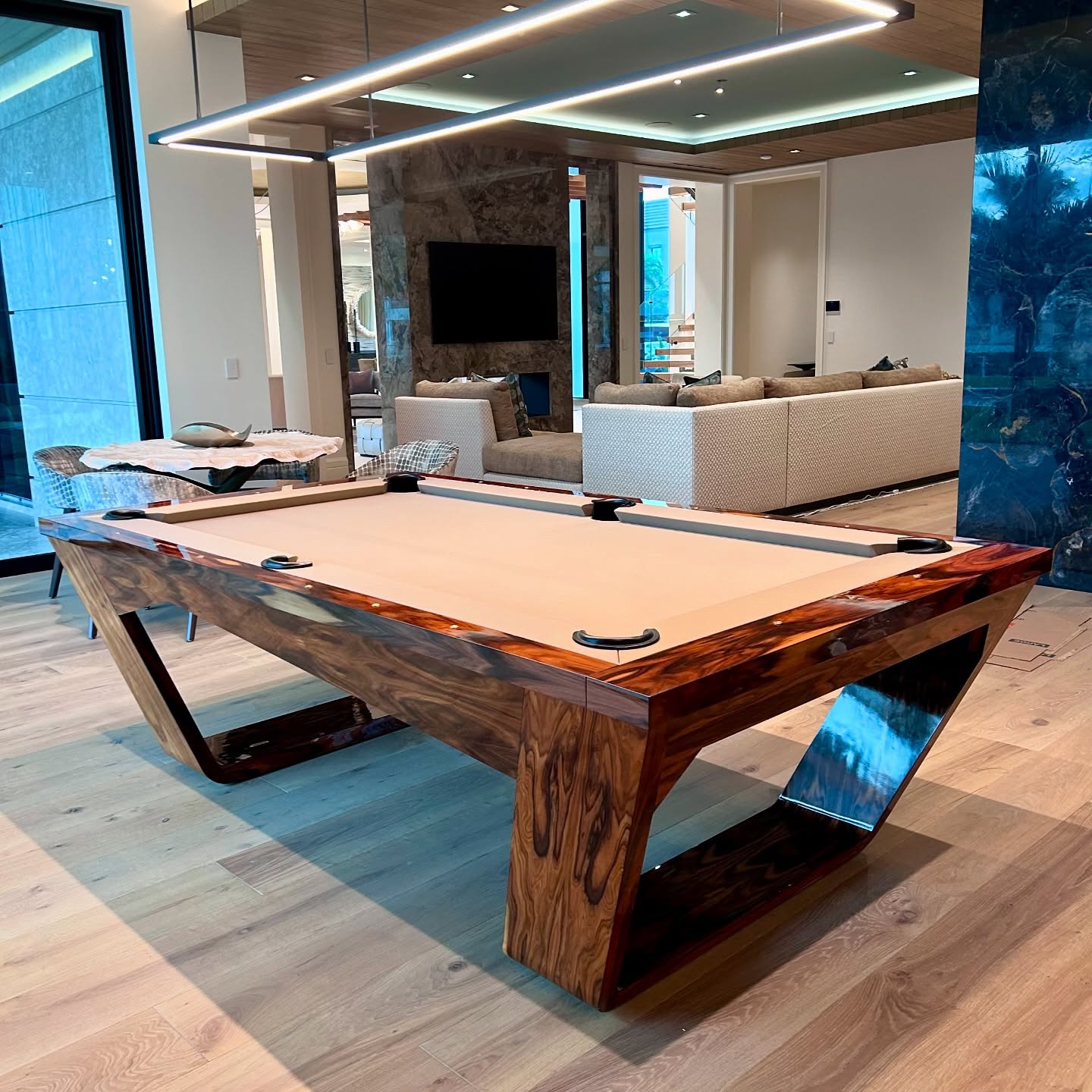 A modern, wooden pool table with a beige playing surface stands in a stylish, open living space with light wood floors, neutral furniture, and large windows overlooking an outdoor area.