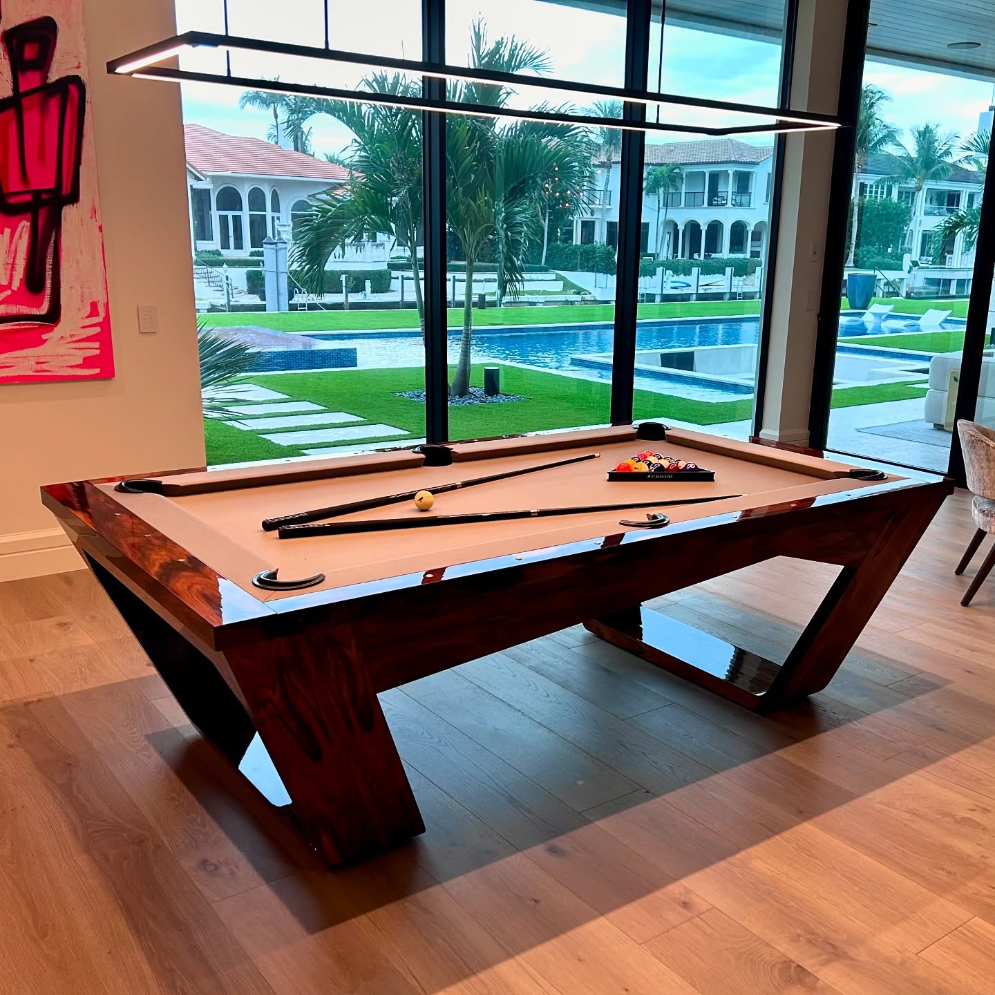 A modern pool table with a wooden frame sits in a stylish room with large windows overlooking a pool, palm trees, and a luxurious house. Pool cues, balls, and a triangle are arranged on the table.