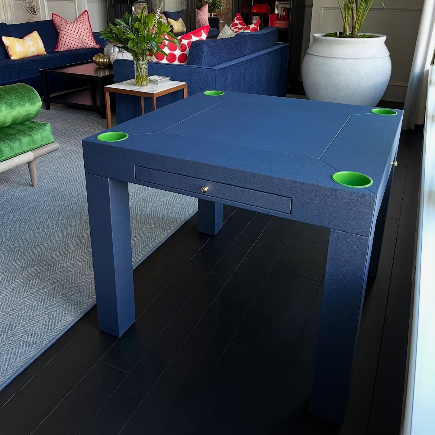 A modern blue square table with green cup holders at each corner stands on a dark wooden floor in a stylish living room with colorful sofas, cushions, plants, and decor.