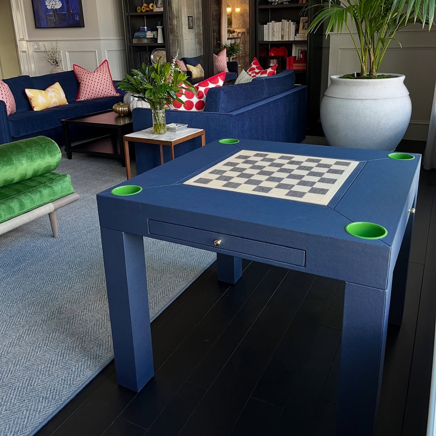 A modern living room features a blue square game table with a chessboard and green cup holders, surrounded by colorful sofas, plants, and decorative pillows.