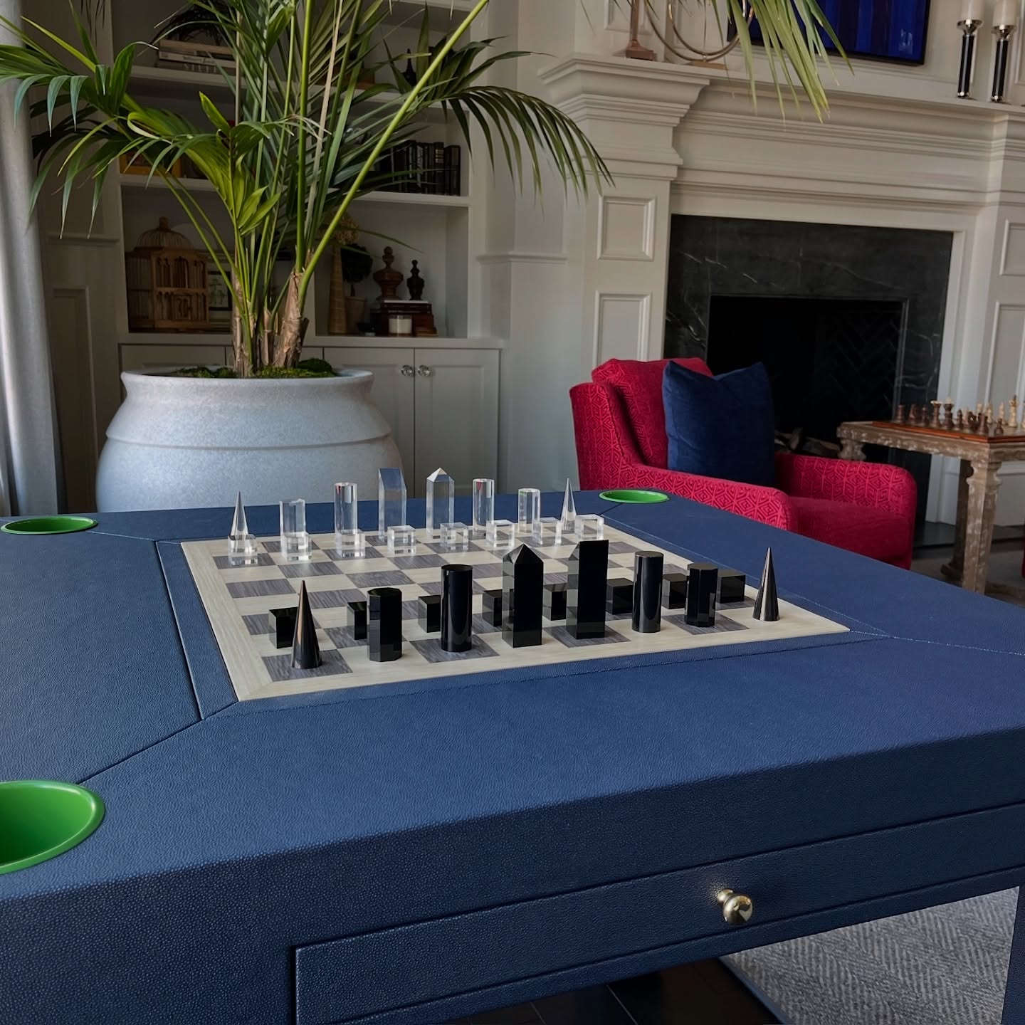 A modern chess set with tall, black and clear geometric pieces is displayed on a blue game table. Behind, there is a large plant, built-in shelves, a fireplace, and a red armchair. Another chess set sits in the background.