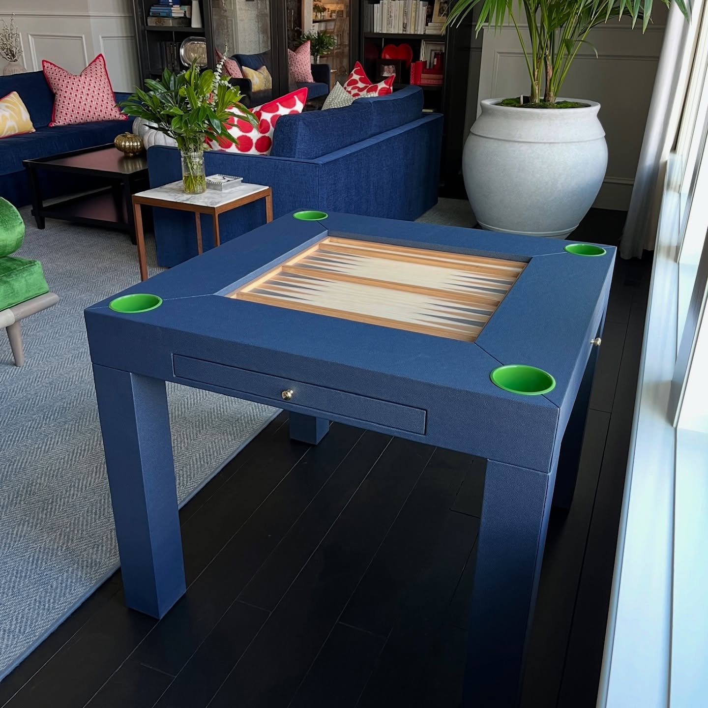 A modern blue square game table with a backgammon board in the center and four green cup holders stands in a stylish living room with blue sofas, a large potted plant, and colorful pillows.