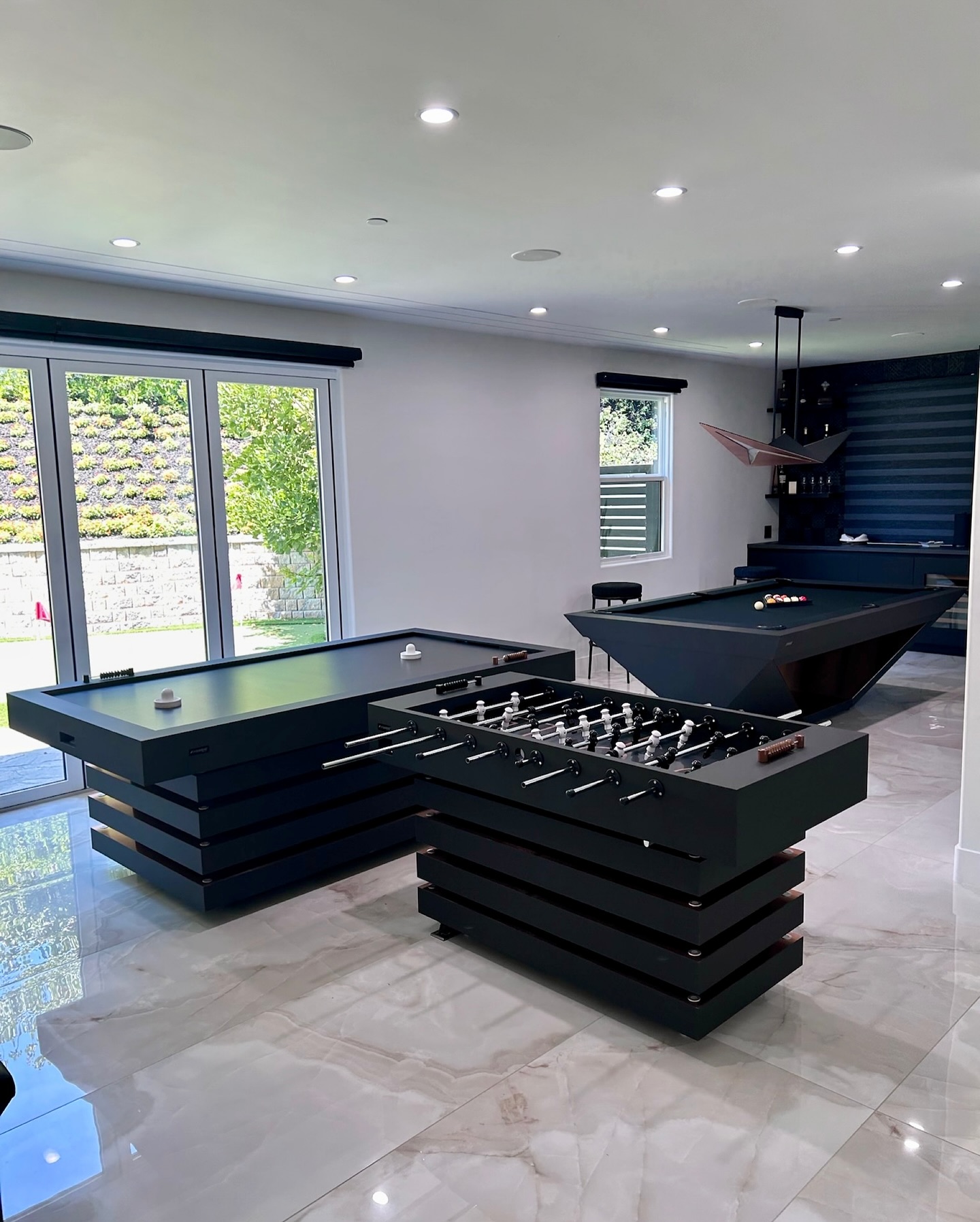 Modern game room with air hockey, foosball, and pool tables on marble flooring, large windows letting in natural light, and a view of greenery outside.
