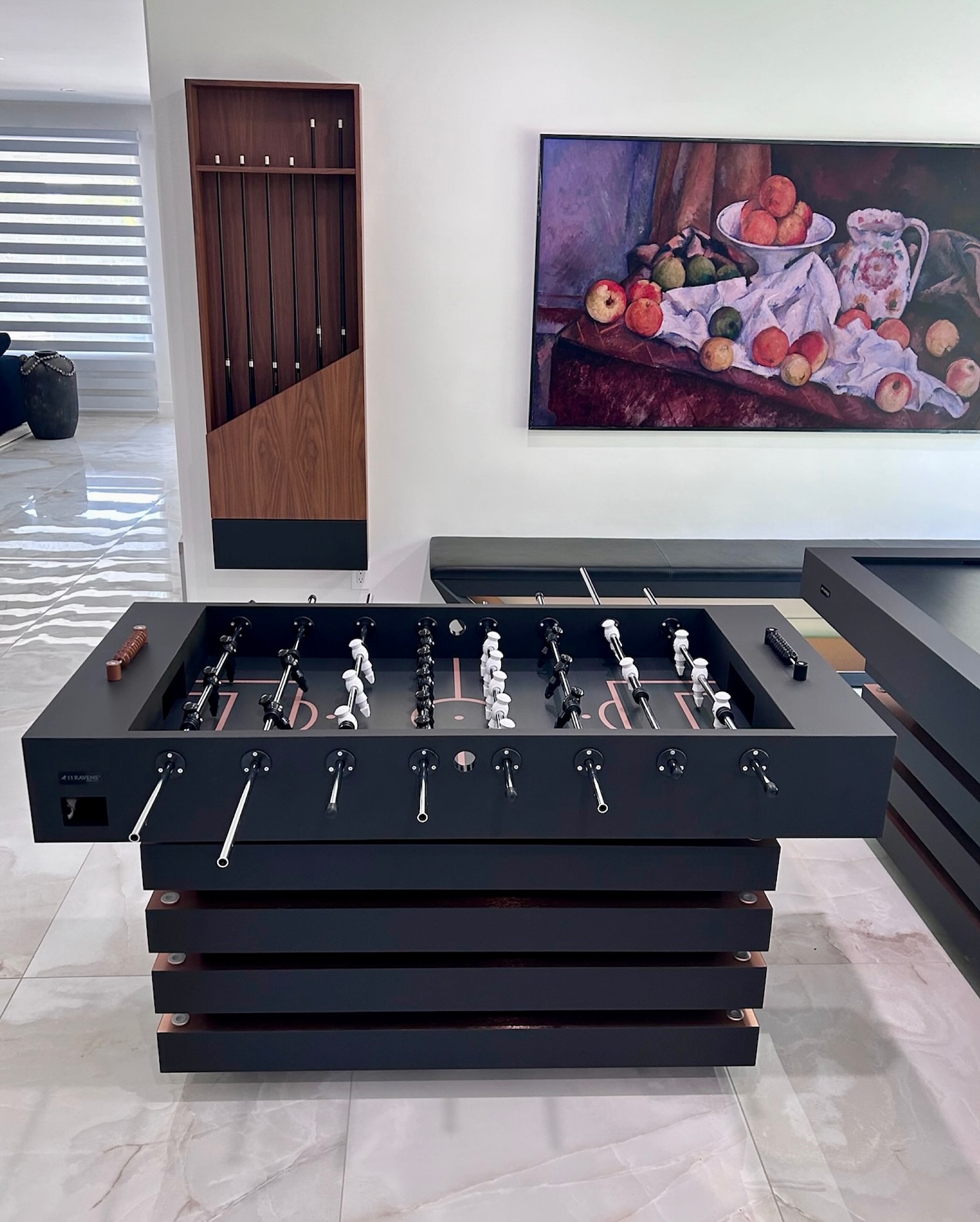 A modern black foosball table stands on a glossy tiled floor in a stylish room with a fruit still life painting on the wall and a wood coat rack in the background.