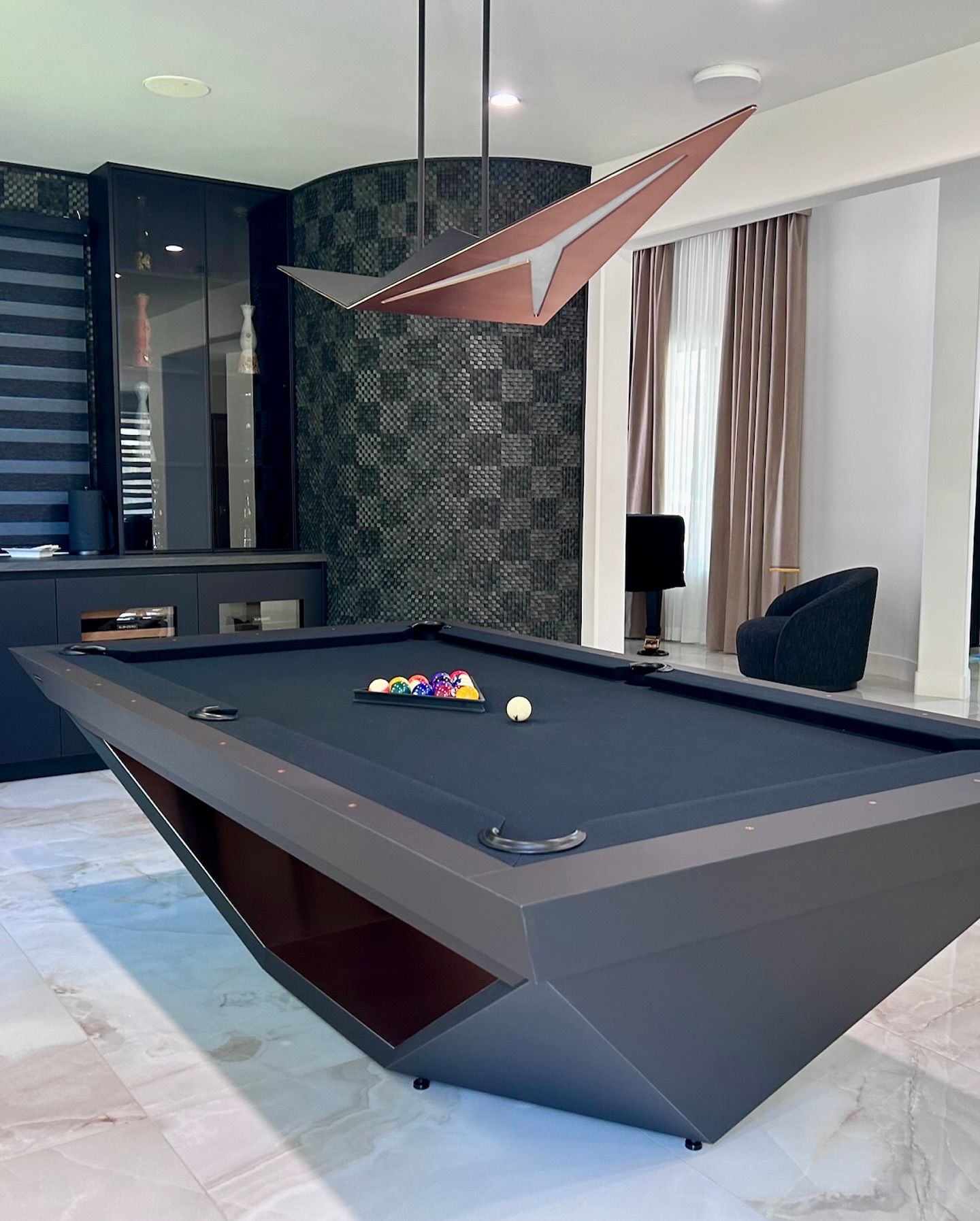 A modern room with a geometric black pool table, a set of billiard balls racked, a cue ball, a sculptural overhead light, marble flooring, and contemporary furniture in the background.