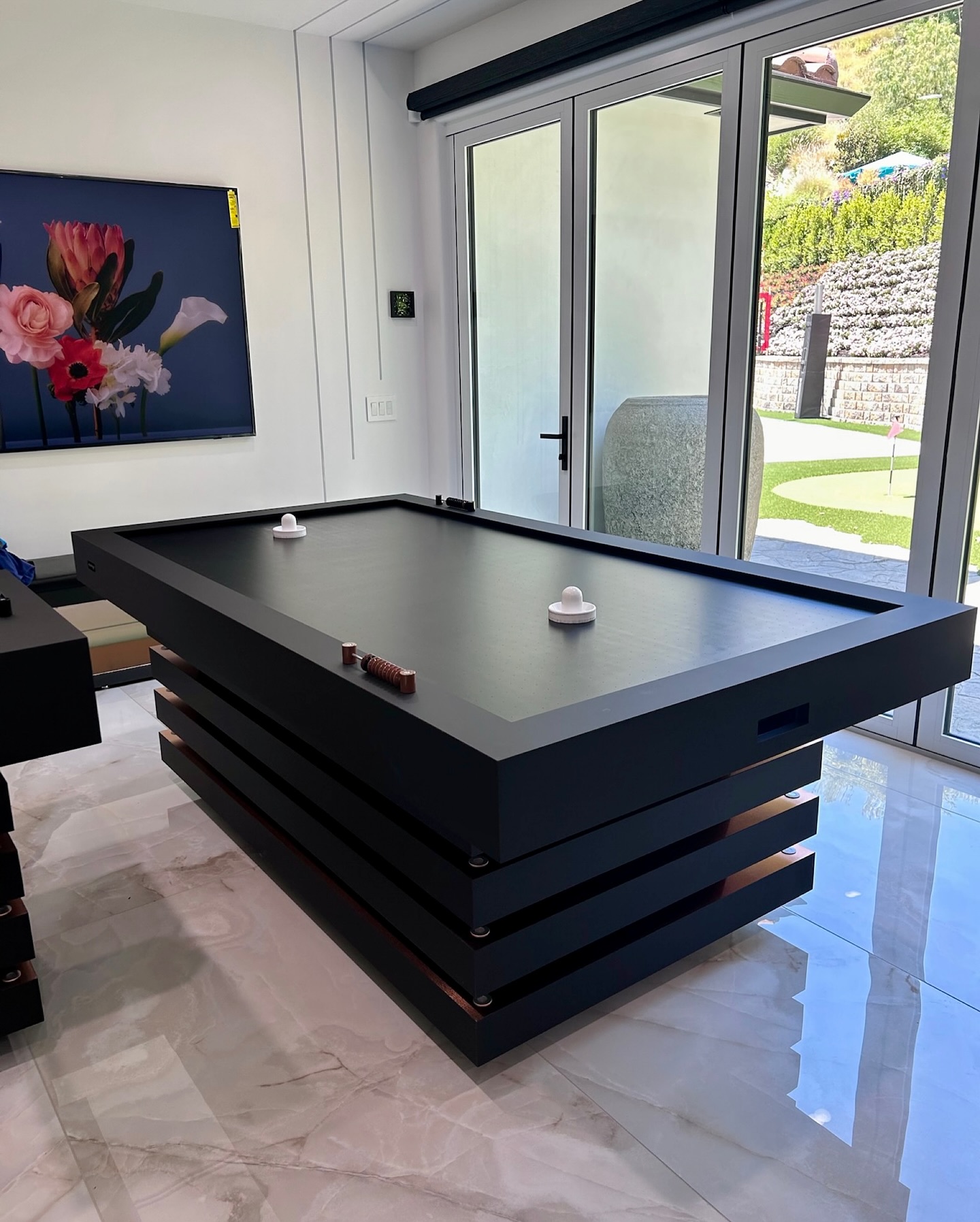 A modern black air hockey table sits in a bright room with large windows. Flower artwork hangs on the wall, and a well-maintained garden and putting green are visible outside through the glass doors.