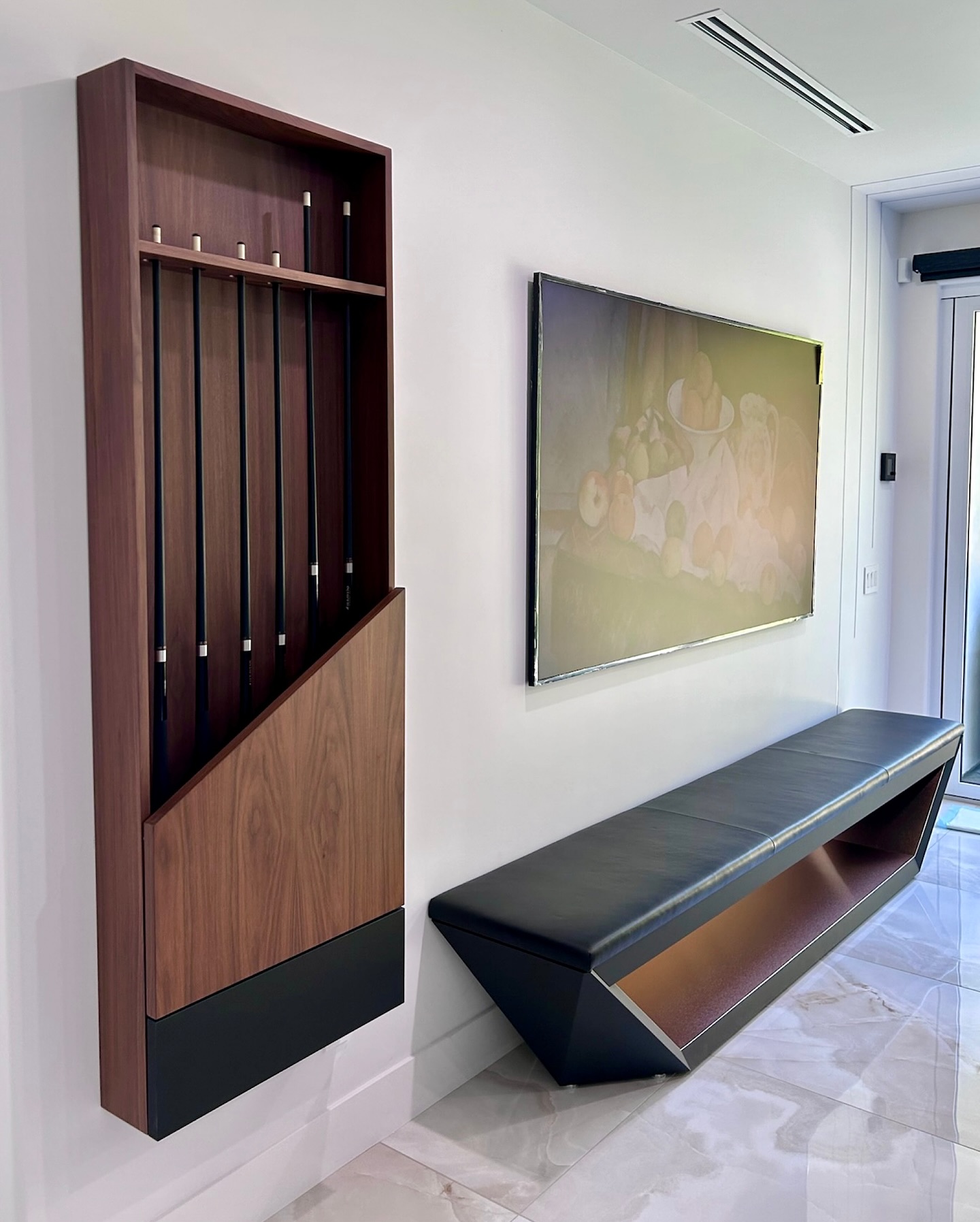 A modern hallway features a wooden cue rack holding five pool cues, a long black cushioned bench with a geometric base, and a framed still life painting on a white wall, with sunlight streaming in from a glass door.