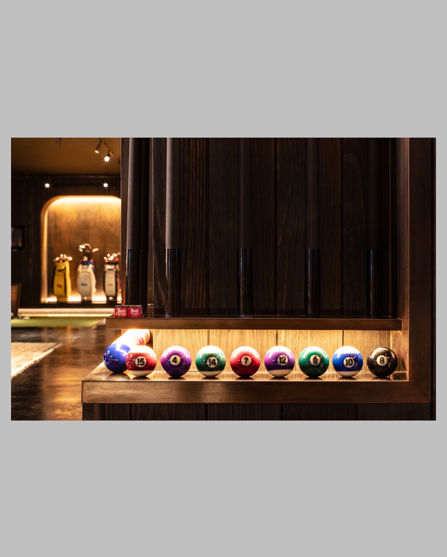 A row of billiard balls numbered 15 through 8 is displayed on a wooden shelf, with golf bags visible in the dimly lit background.