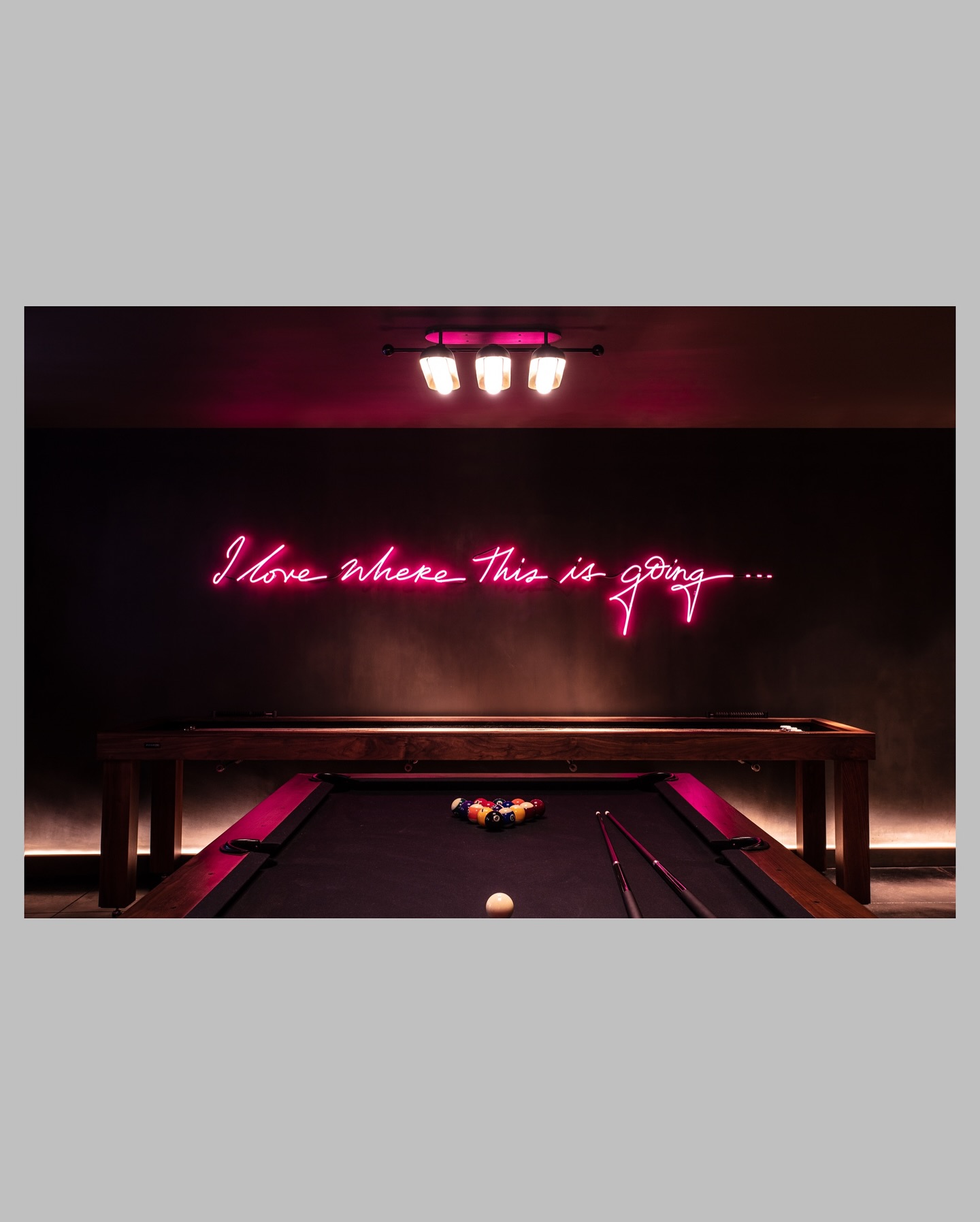 A dimly lit room with a pool table and a pink neon sign on the wall that reads, I love where this is going... Pool balls are racked on the table, with cues resting on its edge.