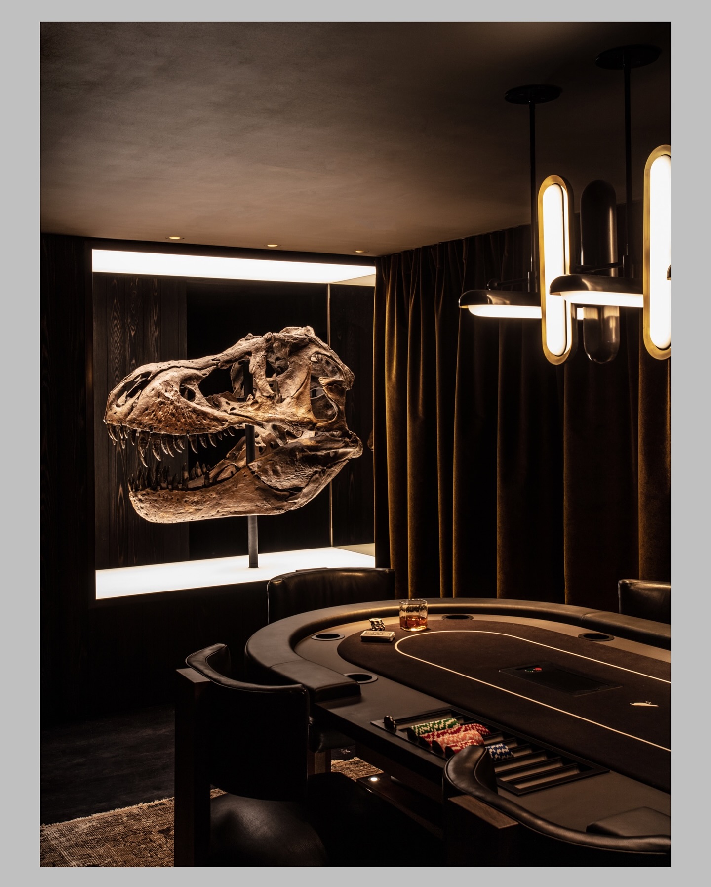 A dimly lit poker room with a round table, poker chips, and drinks, featuring a large, illuminated dinosaur skull displayed on the wall behind the table.