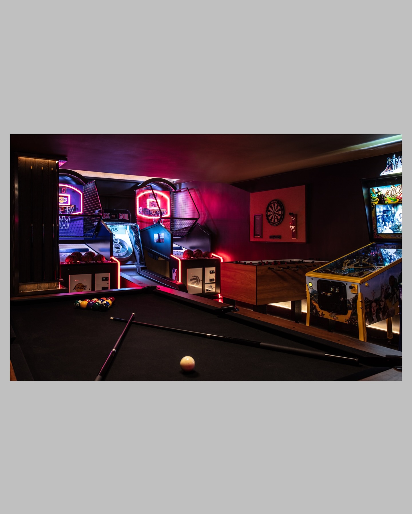 A game room with a black pool table, arcade basketball machines, a foosball table, a pinball machine, and a dartboard, all illuminated by colorful neon lights.
