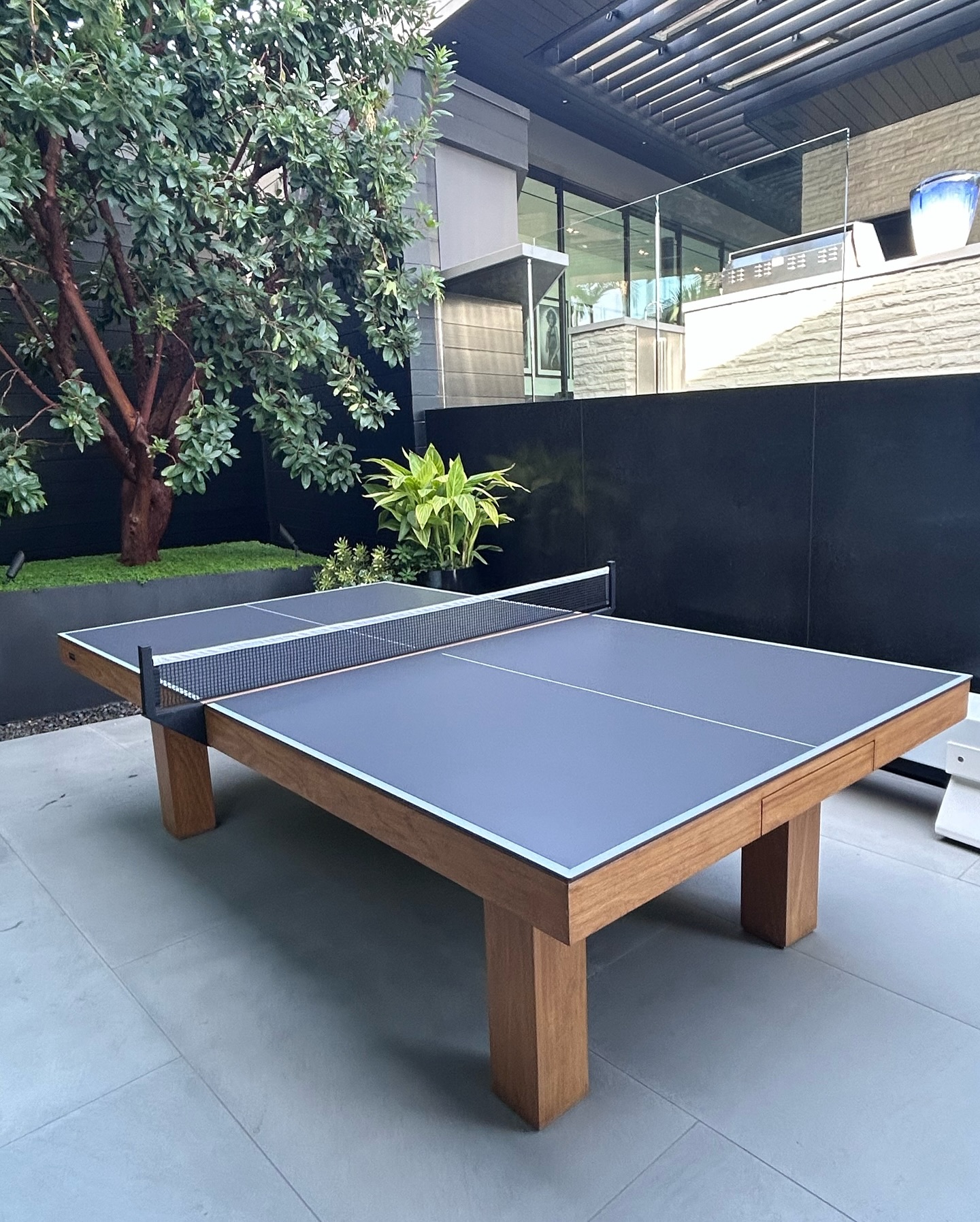 A modern outdoor ping pong table with a wooden frame and blue playing surface is set on a patio near plants and a tree, surrounded by glass and black walls.