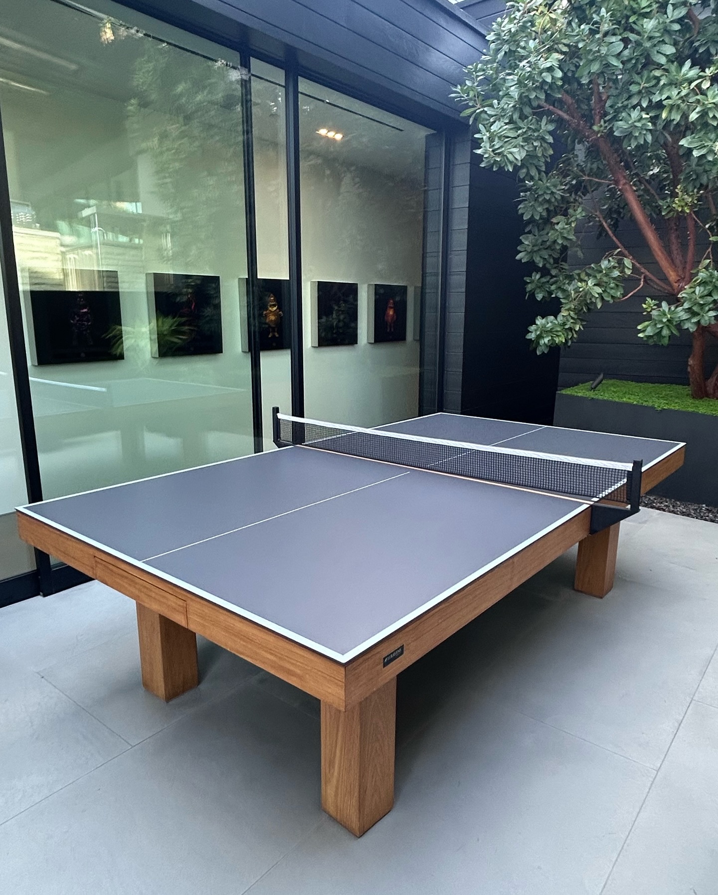 A modern ping pong table with a wooden frame and a gray playing surface is set up next to large glass windows, with a tree and dark walls in the background.
