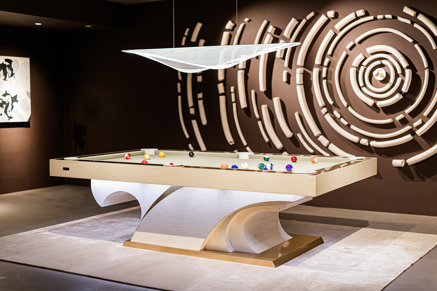 A modern, sculptural pool table with a unique curved base stands on a rug in a stylish room. The walls feature abstract brown and white geometric art, and a sleek, contemporary light fixture hangs above the table.