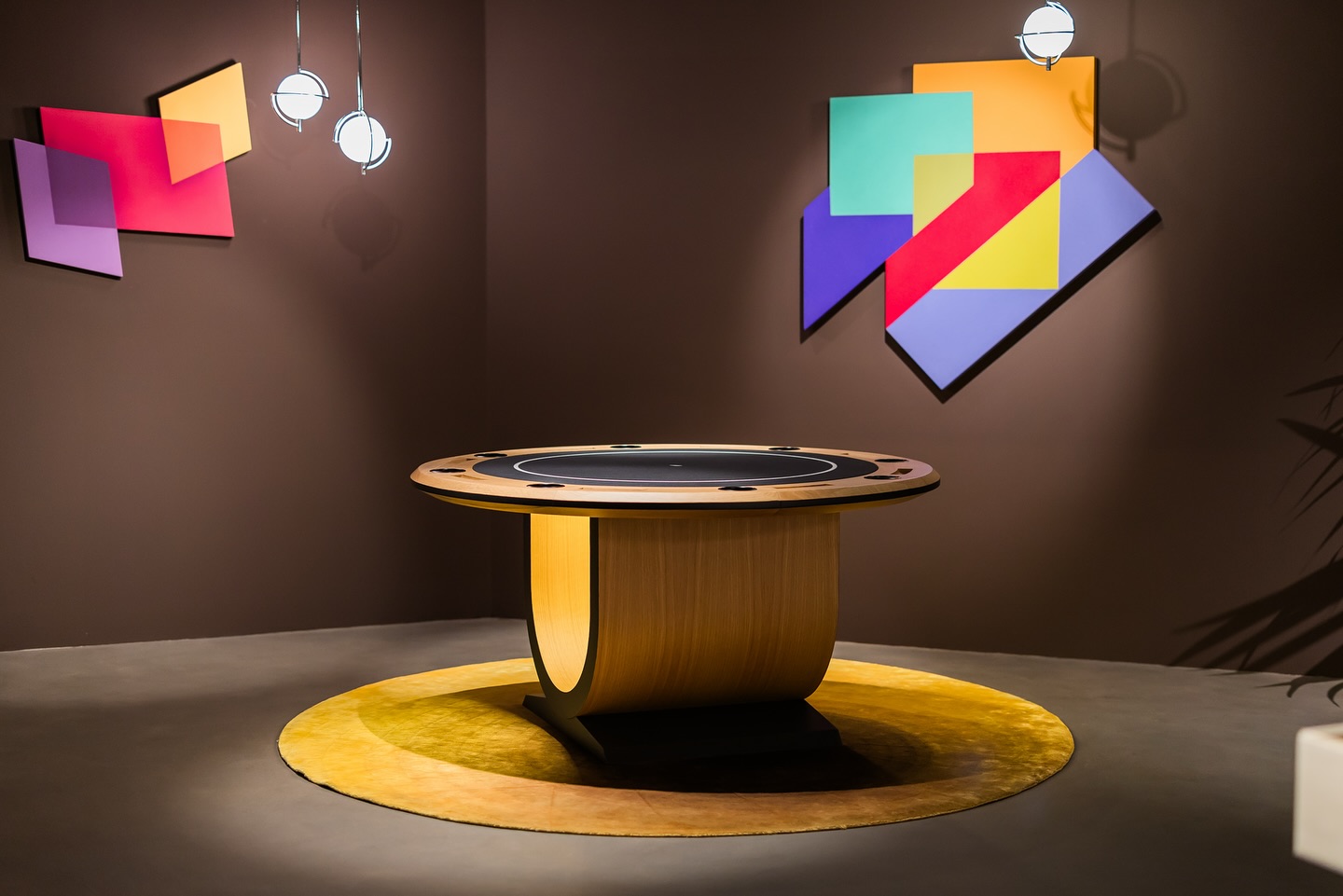 A modern wooden poker table on a round yellow rug, set in a room with brown walls and two colorful abstract geometric art pieces. Two spherical pendant lights hang from the ceiling.