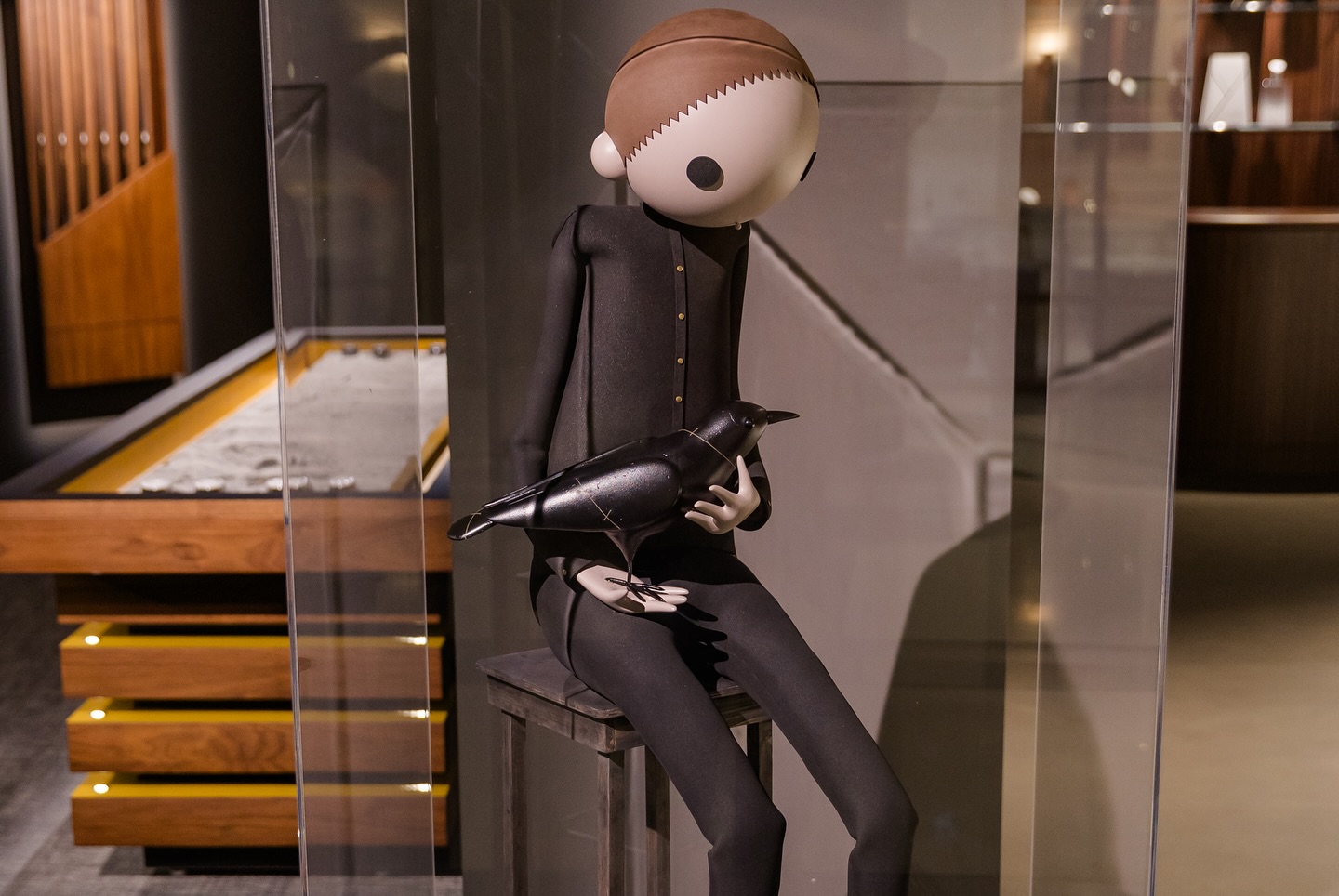 A stylized, faceless human figurine dressed in black sits on a bench, holding a black bird in its lap. The scene is set indoors with wooden furniture and glass panels in the background.