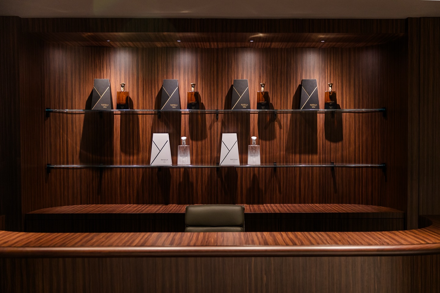 A modern wooden display shelf with several bottles and boxes, lit by spotlights, behind a curved reception desk with a chair. The setting appears luxurious and elegant.