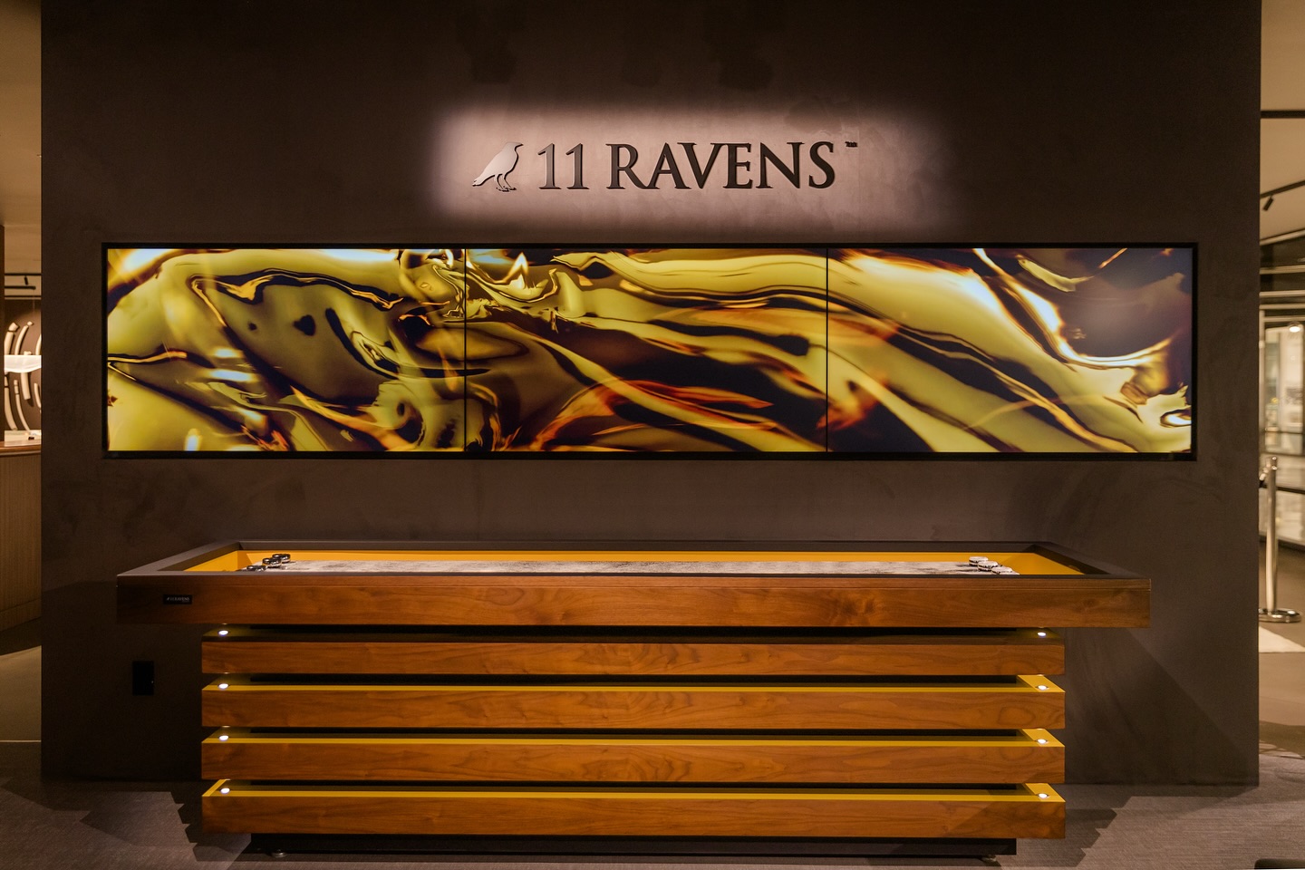 A modern showroom display features a wooden table with horizontal slats below a golden abstract art panel and the 11 Ravens logo on a dark wall.