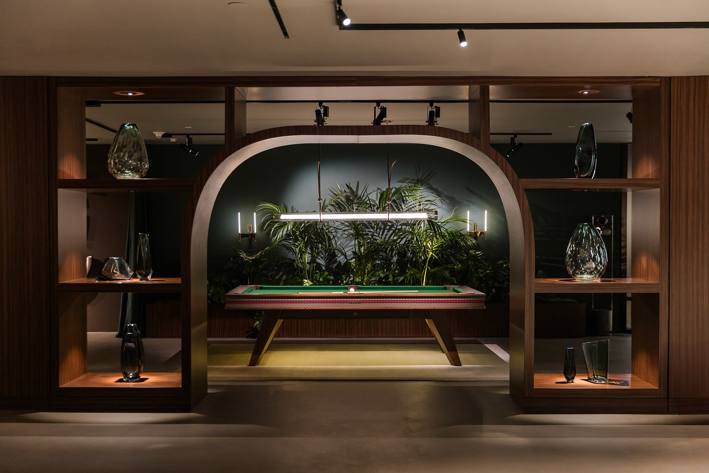 A stylish room with a wooden shelving unit framing a modern pool table. Decorative glass vases sit on the shelves, and lush green plants are visible behind the table under elegant lighting.
