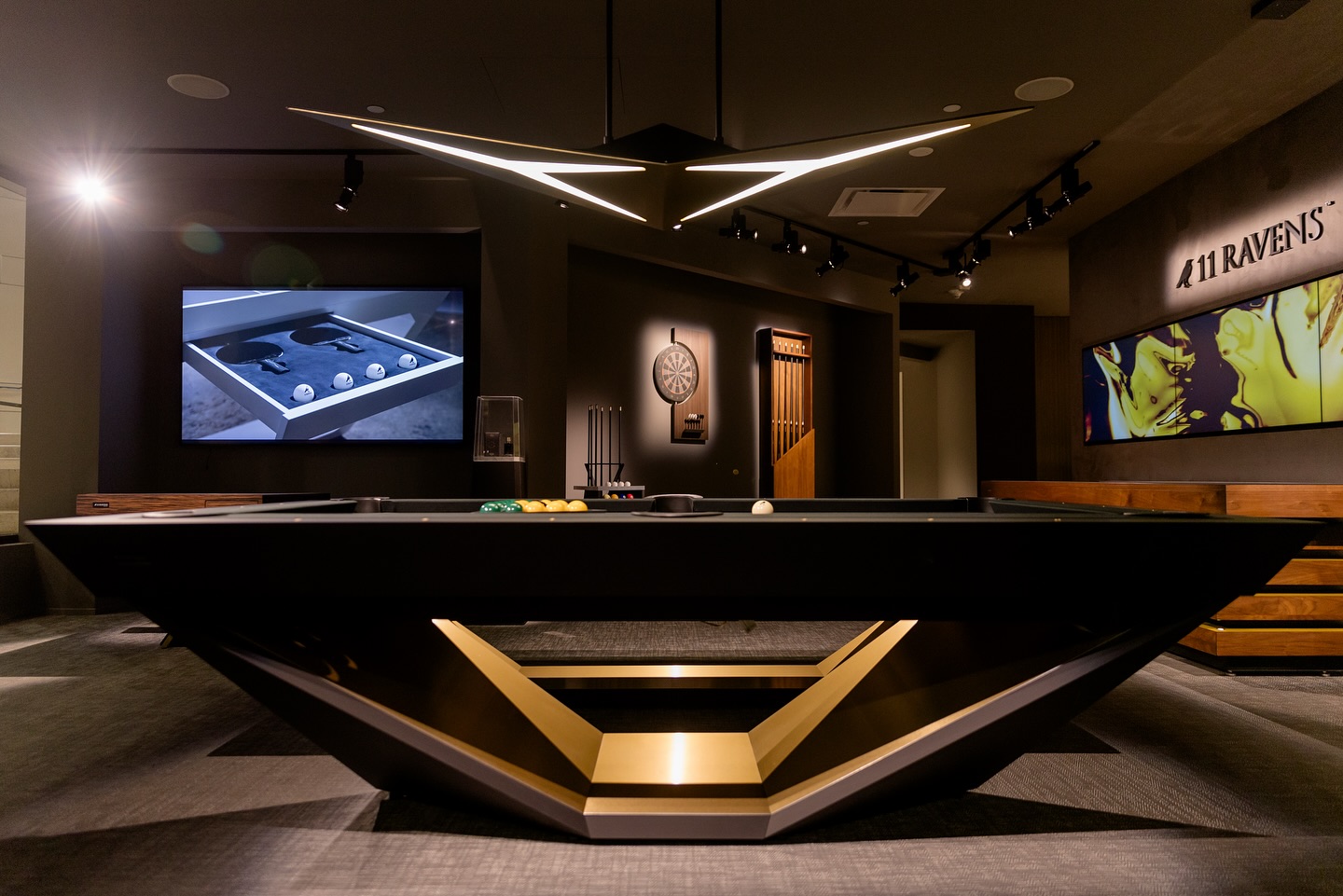 Modern game room with a sleek, geometric black and gold pool table, wall-mounted cues, a dartboard, large TV screen, and contemporary lighting; 11 Ravens sign is visible on the right wall.