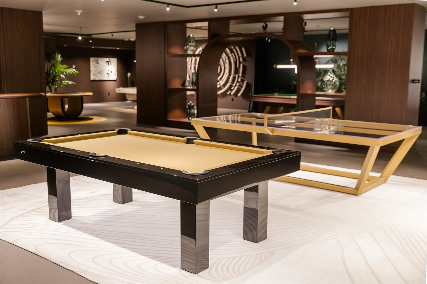Modern game room with a sleek black and beige pool table and a contemporary glass ping pong table, set on a light patterned rug with dark wood walls, soft lighting, and stylish decor in the background.