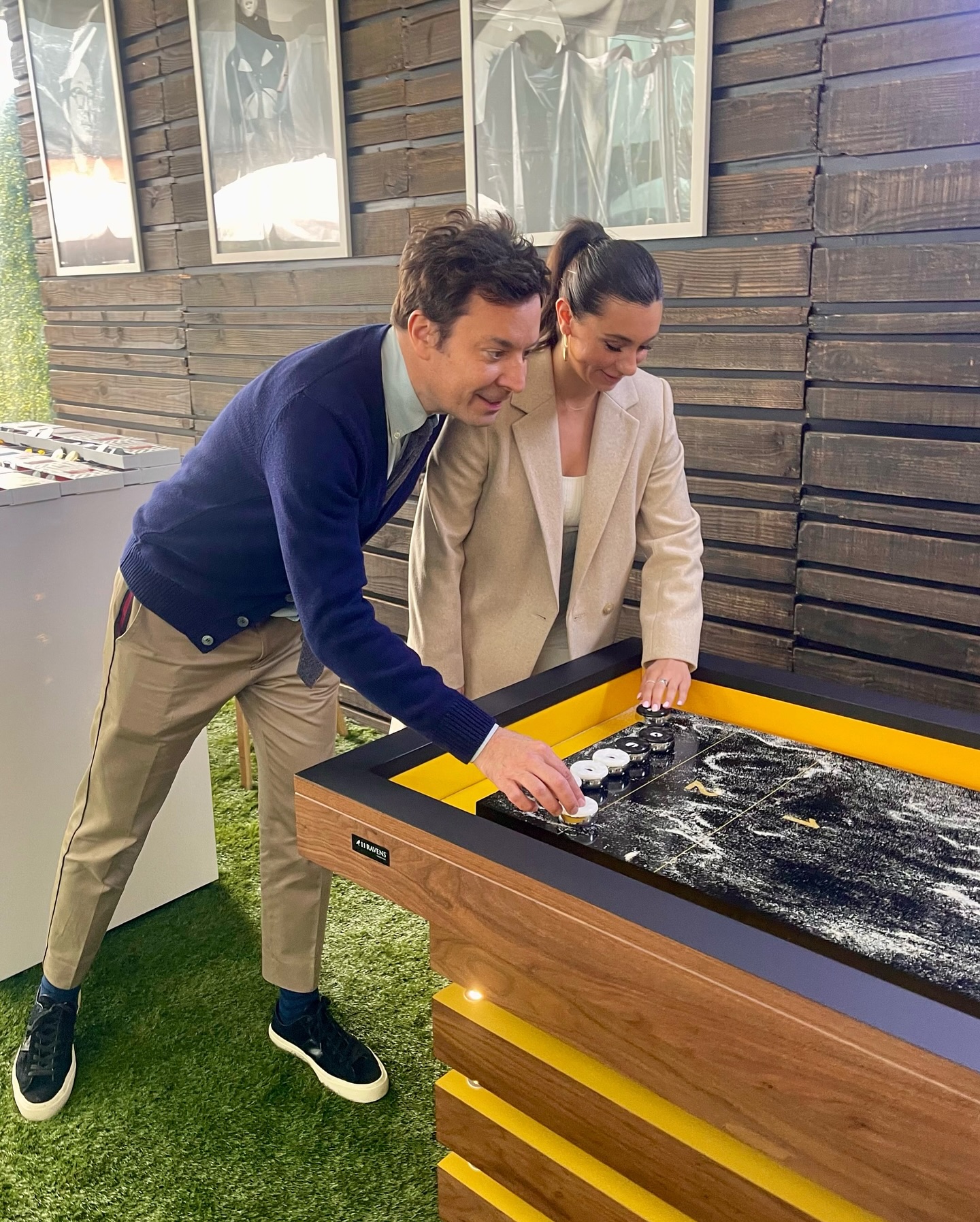 A man and woman smiling as they play shuffleboard together on a wooden tabletop outdoors, standing close to each other, with the man guiding the woman’s hand.