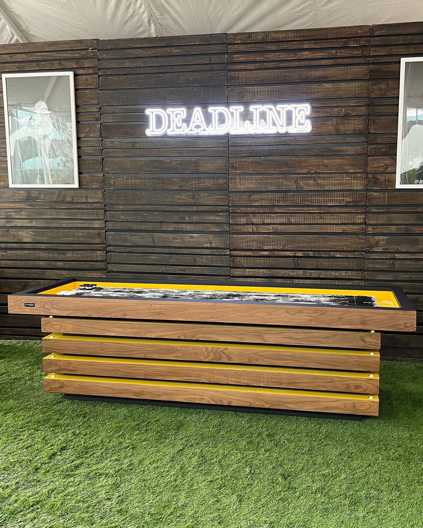 A wooden shuffleboard table with yellow accents sits on green artificial grass in front of a dark wood-paneled wall with framed photos and a neon sign that reads DEADLINE.