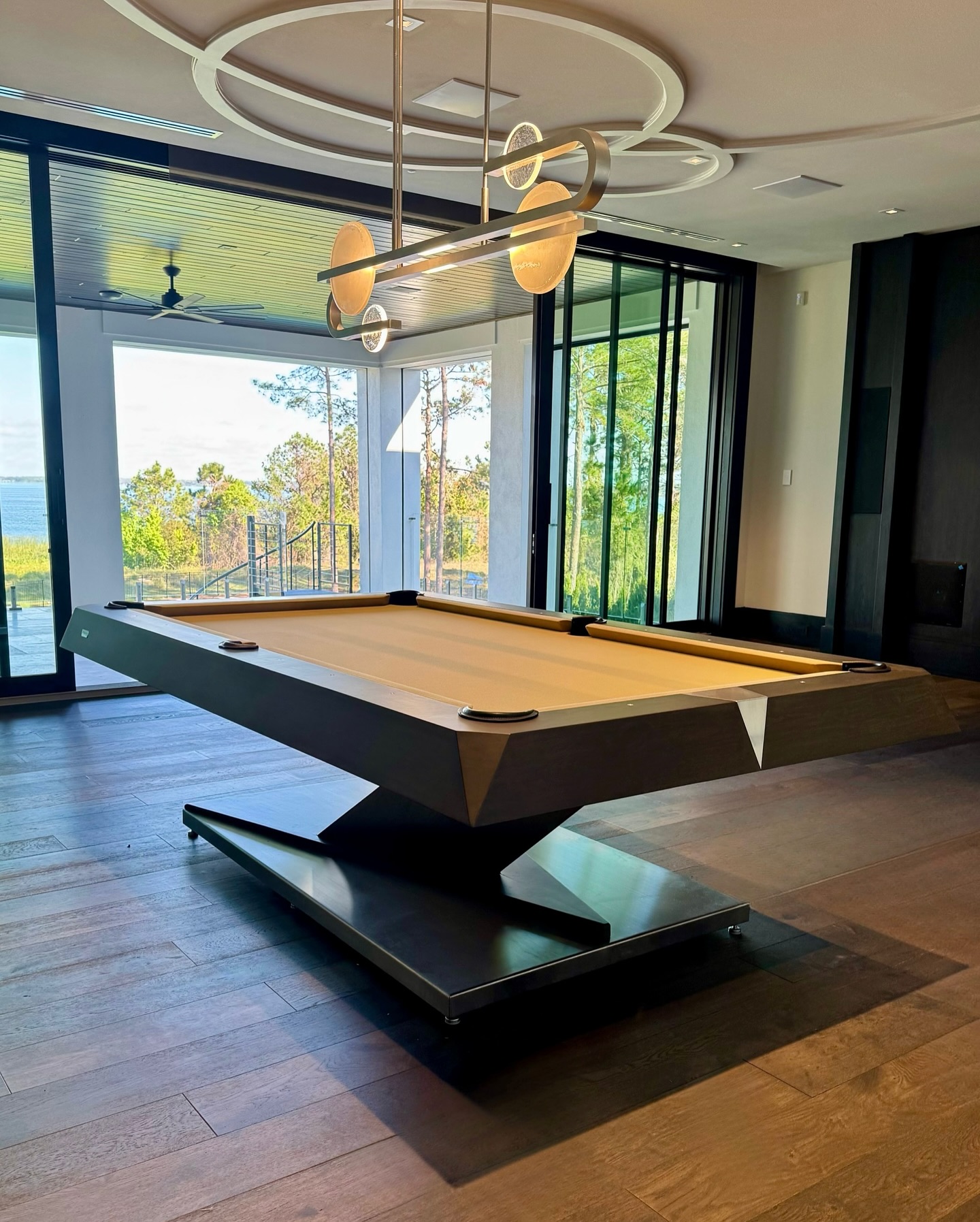 A modern pool table with a unique angular base and tan felt stands in a stylish room with large windows, dark wood floors, and a contemporary light fixture above. Trees and outdoor scenery are visible through the glass.