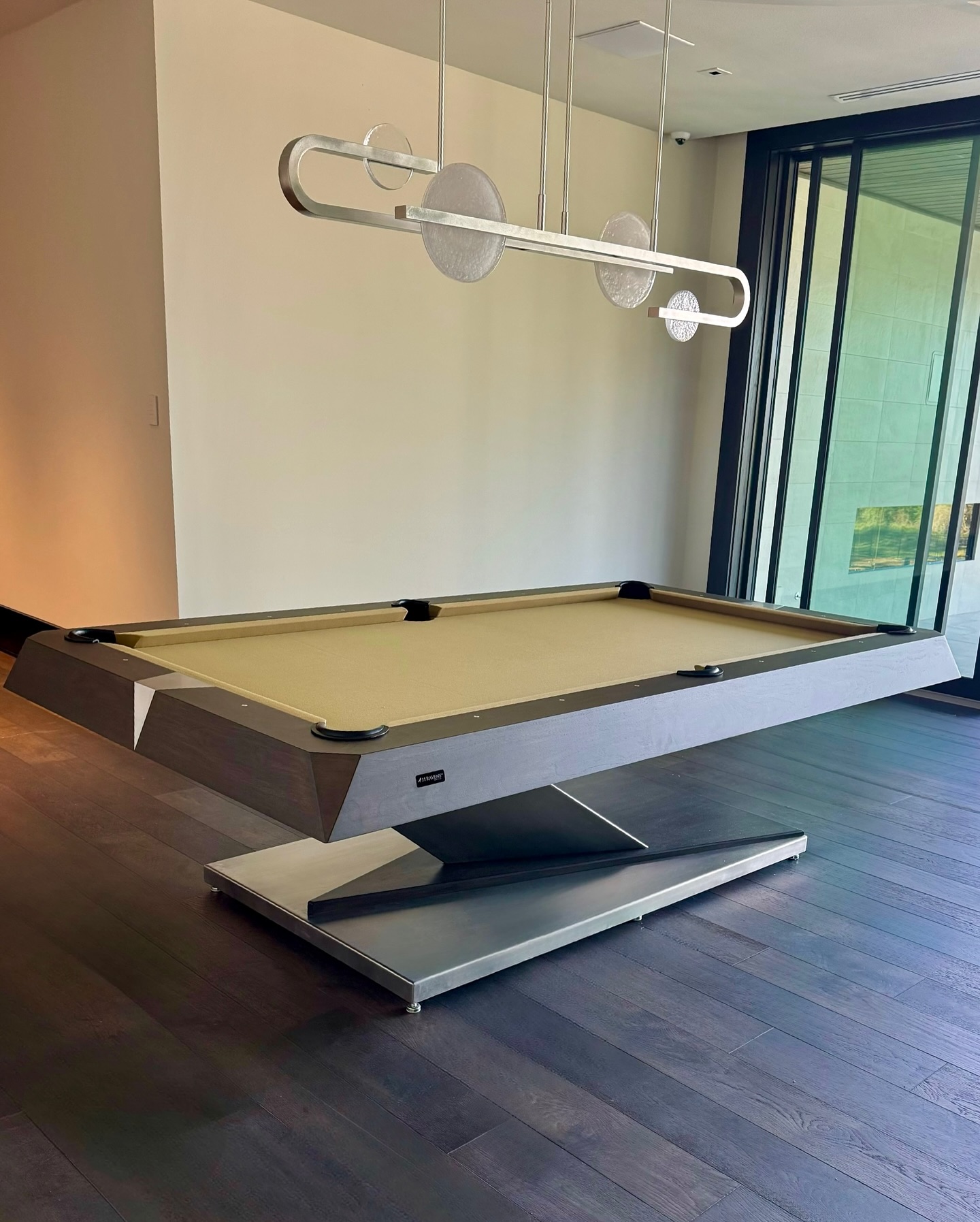 A modern pool table with a sleek metal base and beige felt top sits in a room with dark wood floors and a contemporary light fixture overhead, near large glass sliding doors.