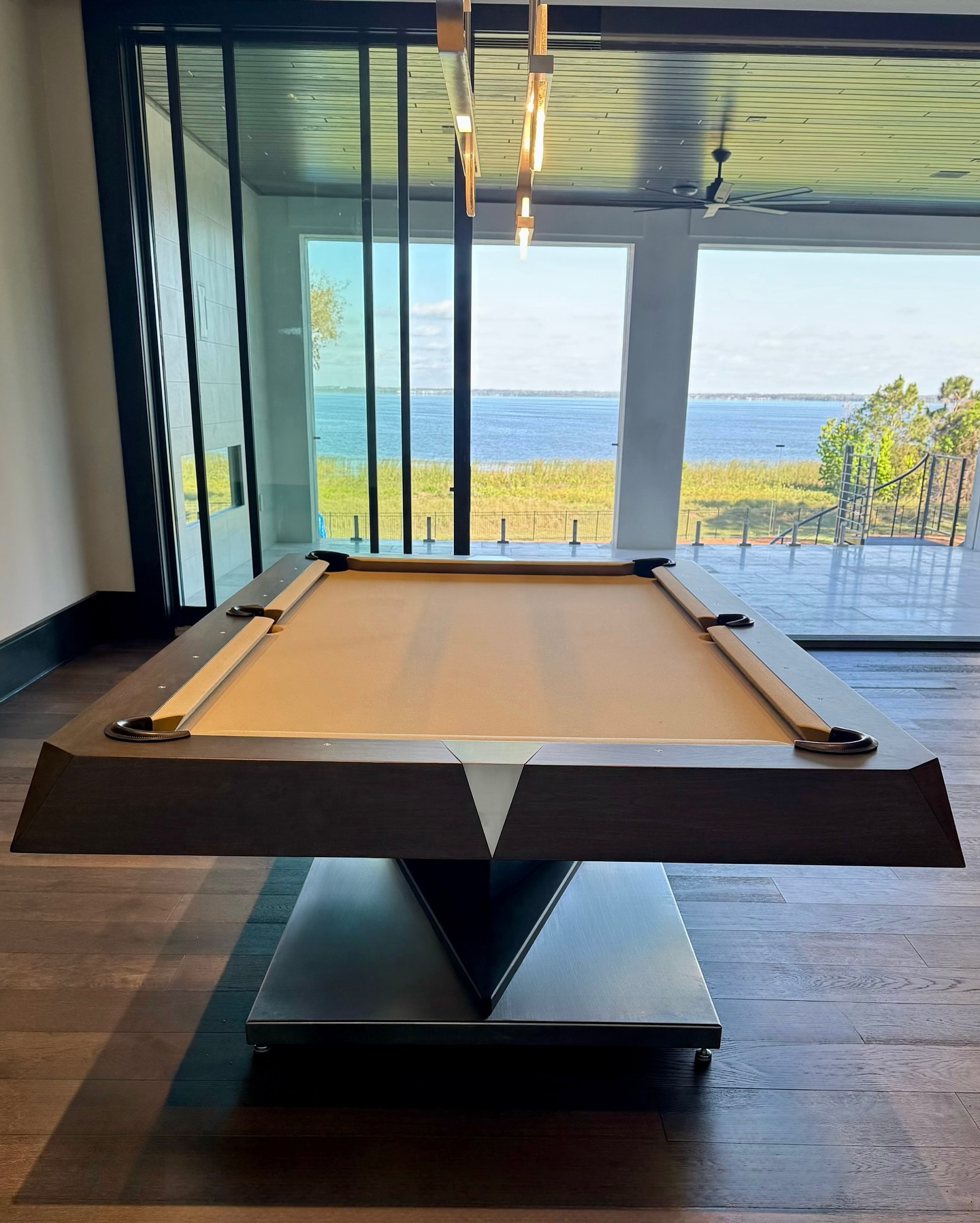 A modern pool table with a tan felt top stands in a room with wood flooring, large glass doors, and a scenic view of a lake and blue sky outside.
