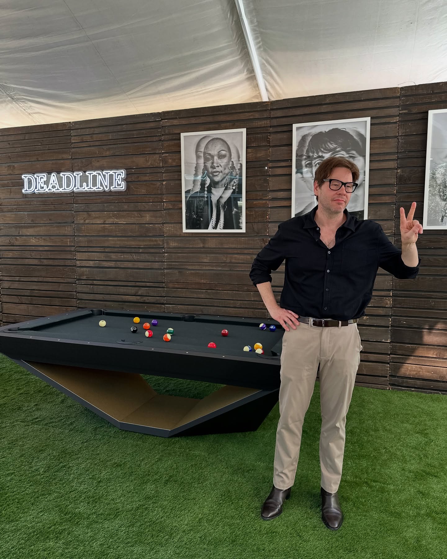 A man in glasses and a black shirt stands by a modern pool table on green artificial grass, flashing a peace sign. Behind him is a wooden wall with black-and-white portraits and a lit DEADLINE sign.
