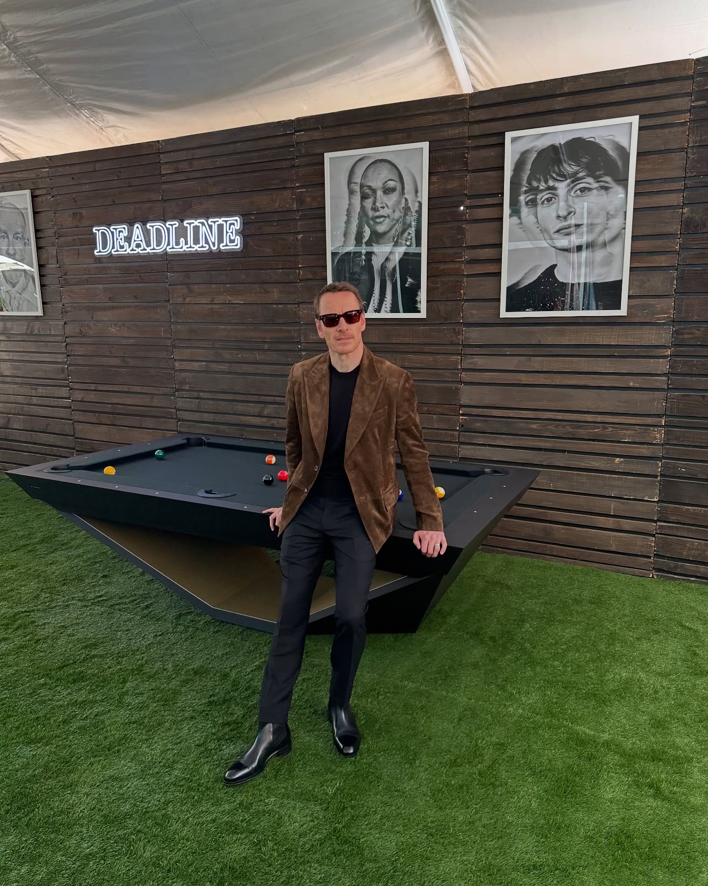 A man in sunglasses, a brown jacket, and black pants stands by a black pool table on green turf under a tent. Behind him, there are Deadline magazine portraits on a wooden wall.