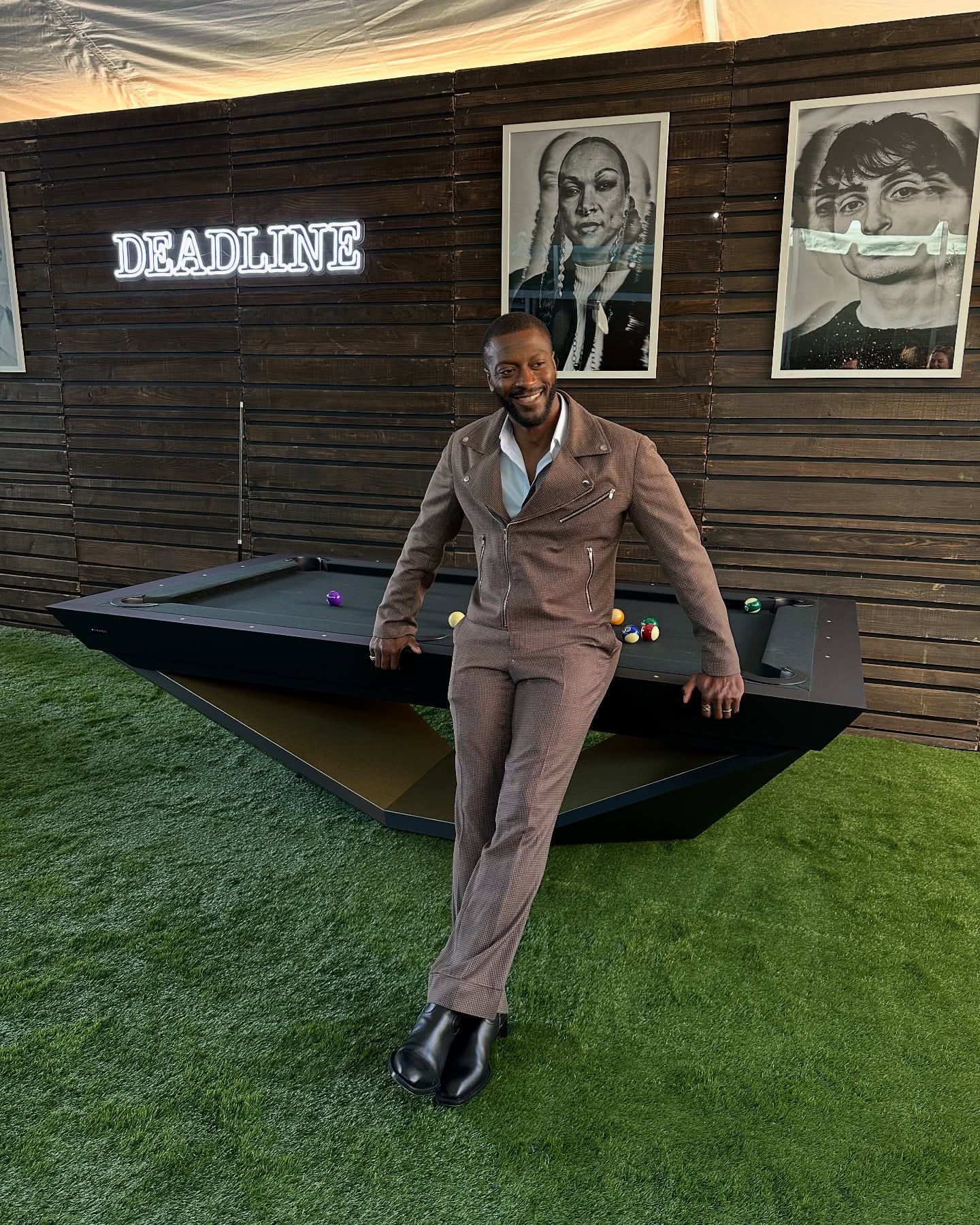 A man in a brown suit leans against a modern black pool table on green turf, smiling. Behind him, a wooden wall displays a DEADLINE neon sign and three large black-and-white portrait photos.