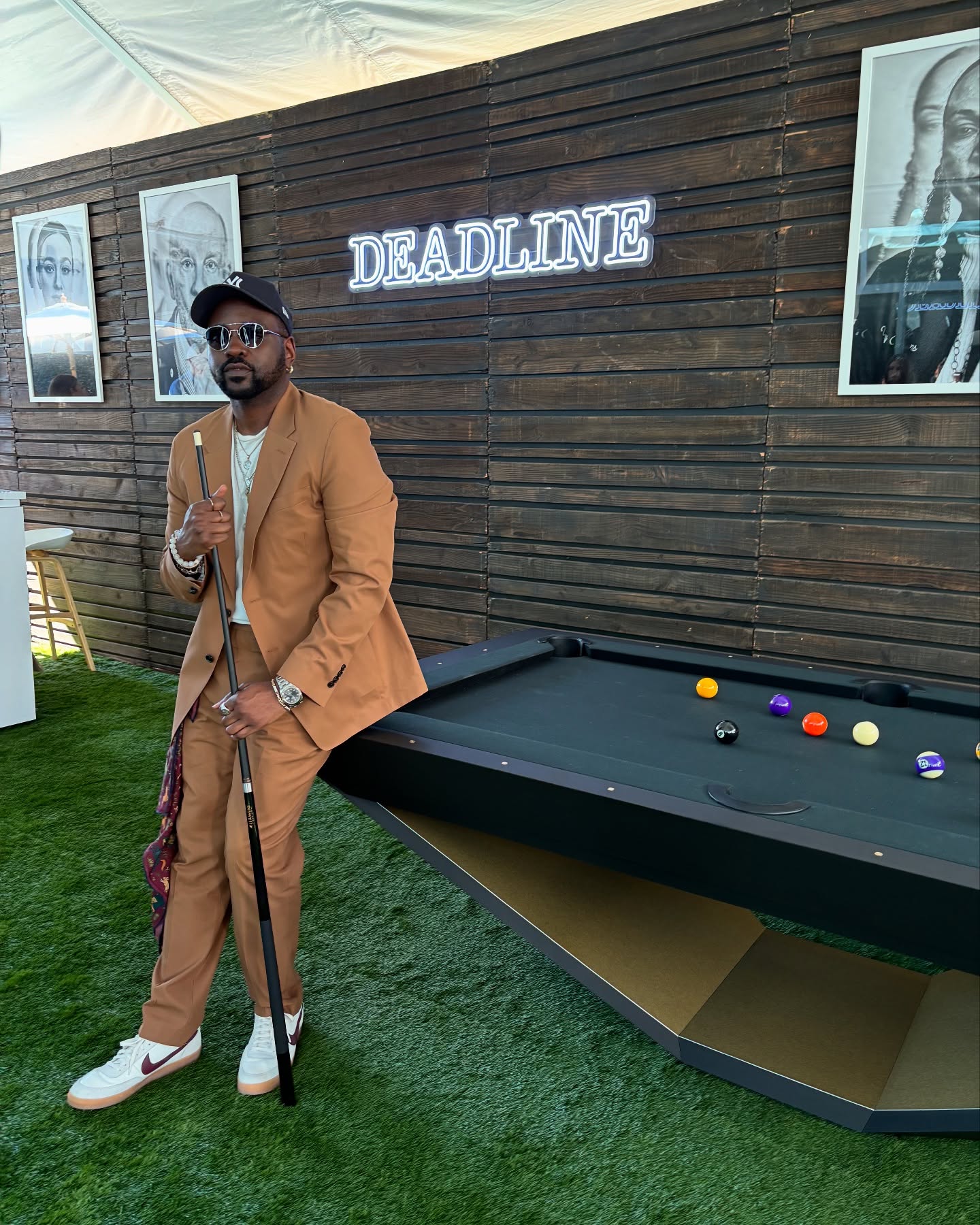 A man in a tan suit and sunglasses stands by a black pool table holding a cue stick. Behind him is a wooden wall with framed photos and a neon “DEADLINE” sign. The floor is covered with green artificial grass.