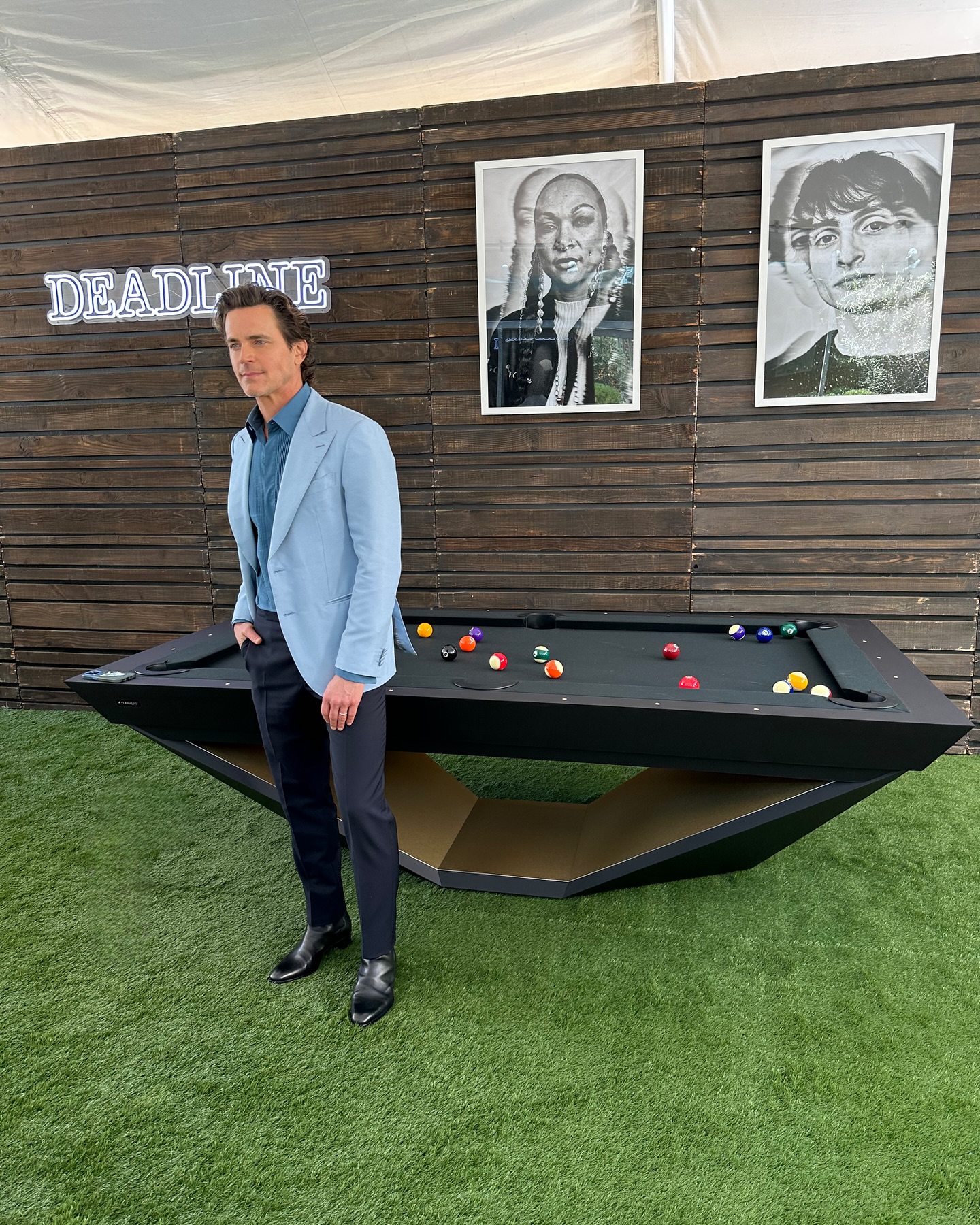 A man in a light blue blazer and dark pants stands beside a modern black pool table on green turf, with wooden paneling, a DEADLINE sign, and two black-and-white portraits in the background.