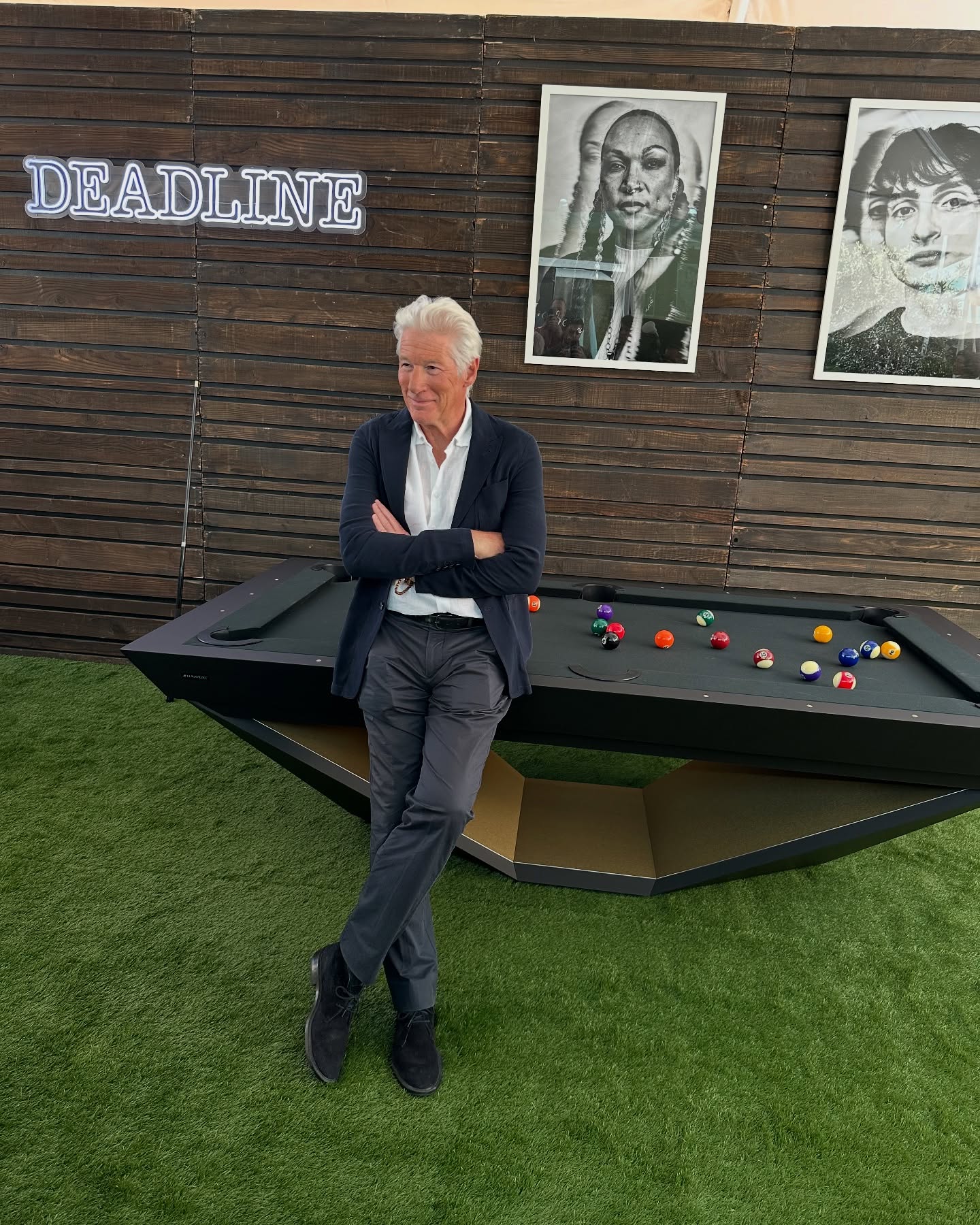 An older man with gray hair, dressed in a dark jacket and pants, stands with arms crossed in front of a black pool table. Behind him are celebrity portraits and a neon DEADLINE sign on a wooden wall. The floor is artificial grass.