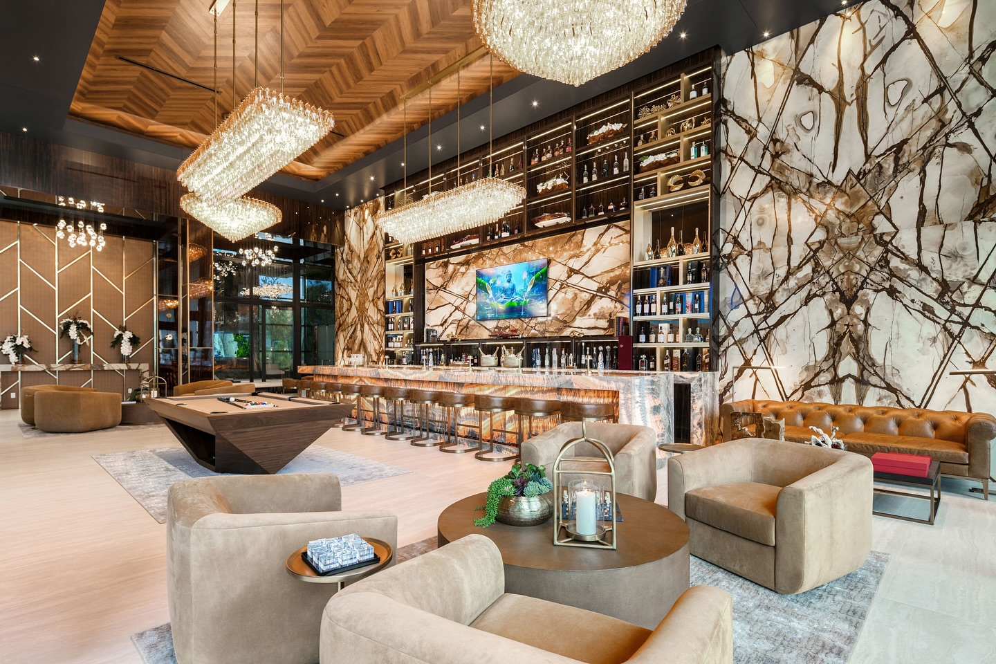Luxurious lounge with plush beige seating, marble walls, a long bar with high stools, grand chandeliers, a pool table, modern decor, and floor-to-ceiling shelves displaying bottles and decorative pieces.