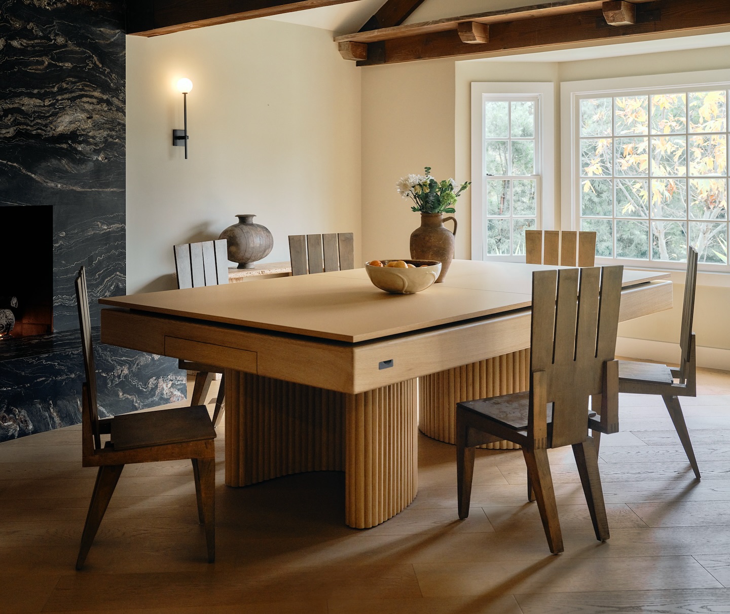 A modern dining room with a wooden table and six matching chairs, a bowl of fruit on the table, large windows letting in natural light, and decorative vases on a sideboard. Exposed beams and stone accents add warmth.