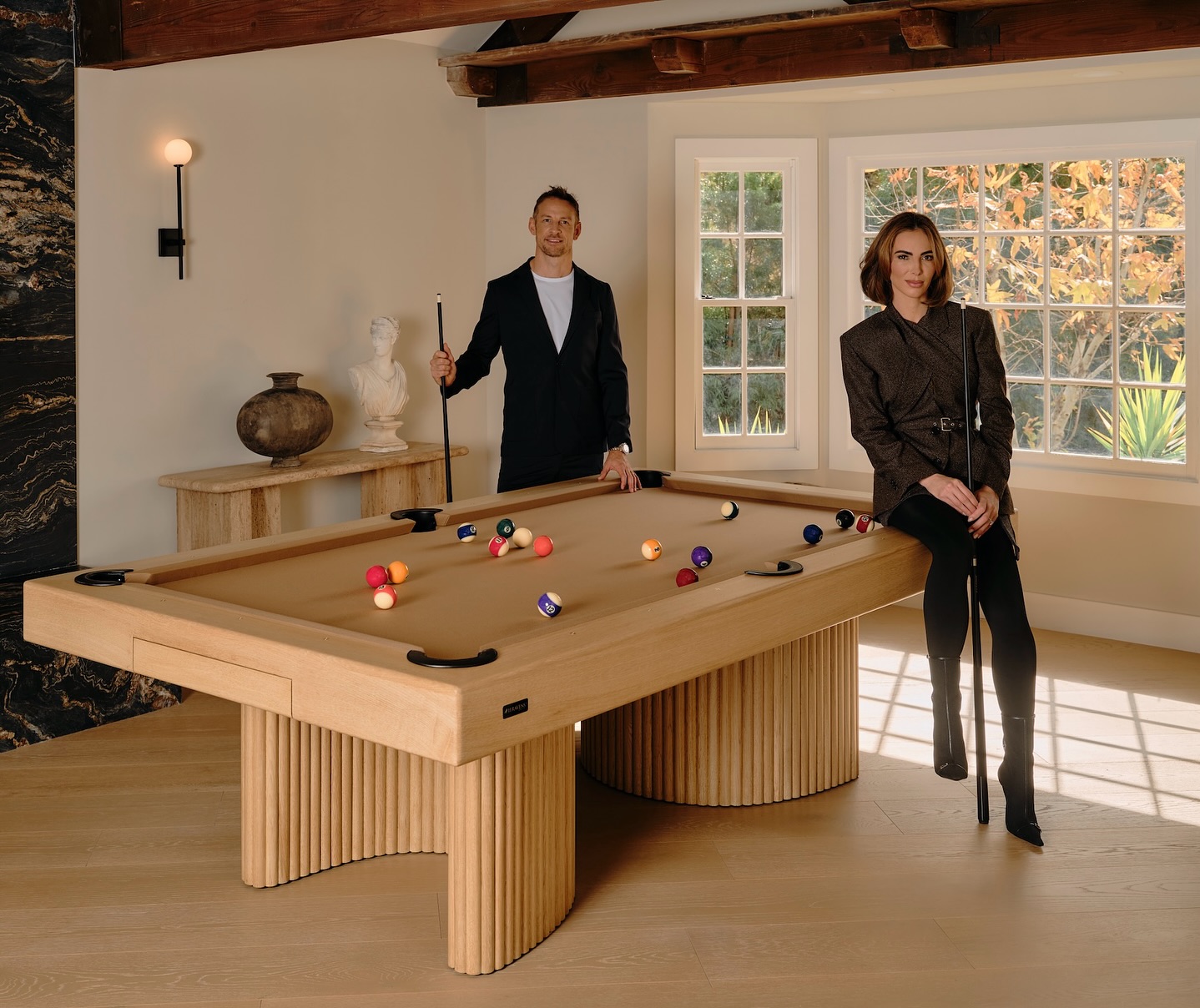 A man holding a pool cue stands beside a tan pool table while a woman sits on its edge. They are in a stylish room with wood floors, a large window, and decorative objects on a side table.