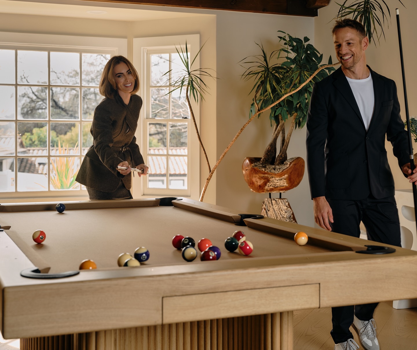 A woman and a man, both dressed in suits, play pool together in a sunlit room with large windows, indoor plants, and a modern wooden pool table. They are smiling and enjoying the game.