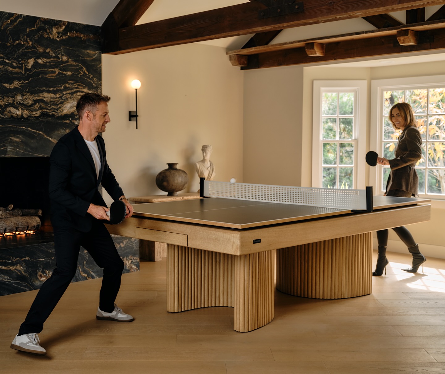 A man and woman play table tennis in a stylish living room with wooden floors, large windows, a modern fireplace, and decorative vases on a side table.