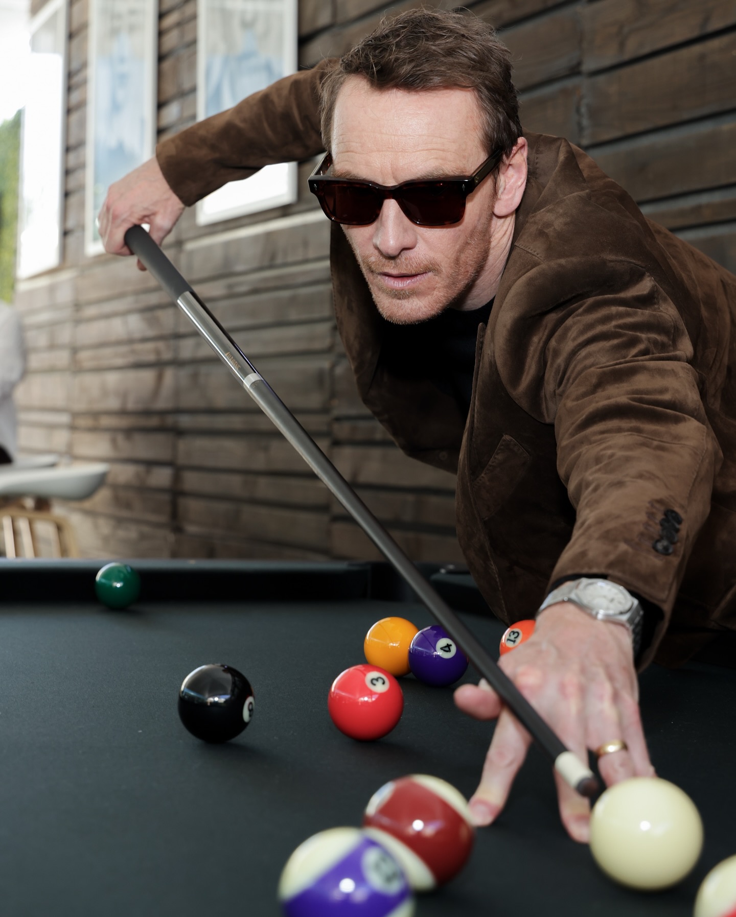A man wearing sunglasses and a brown jacket lines up a shot while playing pool, leaning over the table and aiming his cue at the cue ball. The background features wooden paneling and framed pictures.