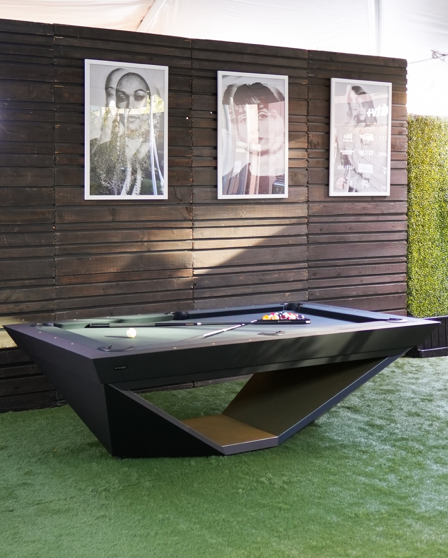 A modern, angular black pool table sits on green artificial grass in front of a wooden slat wall, with three framed black-and-white portraits hanging above it. Pool balls and cues rest on the table.
