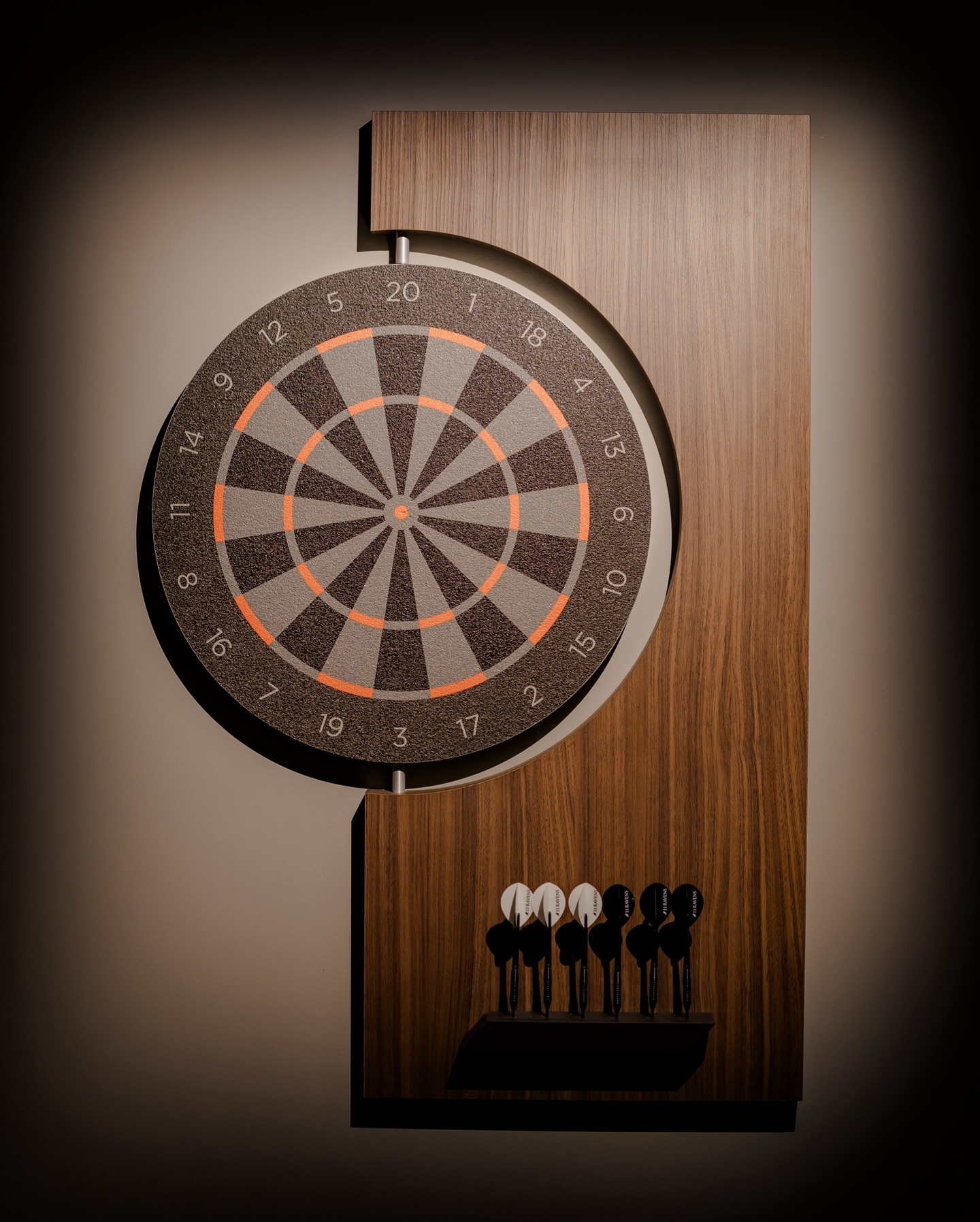 A dartboard mounted on a wooden panel with six darts—three black and three white—placed in a holder below the board, illuminated by a soft light.